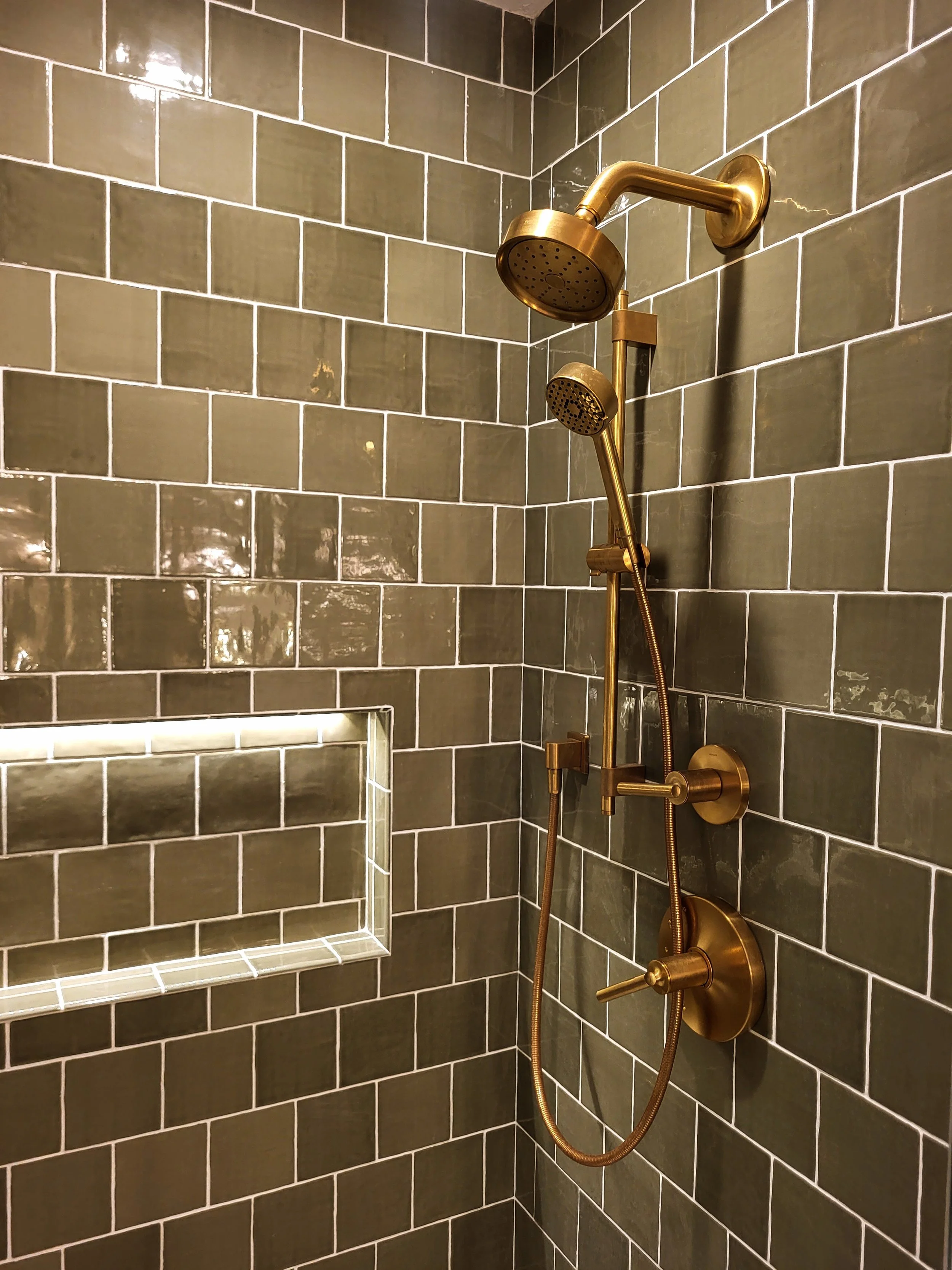 Modern brass shower head against classic green tile next to lighted shower niche.