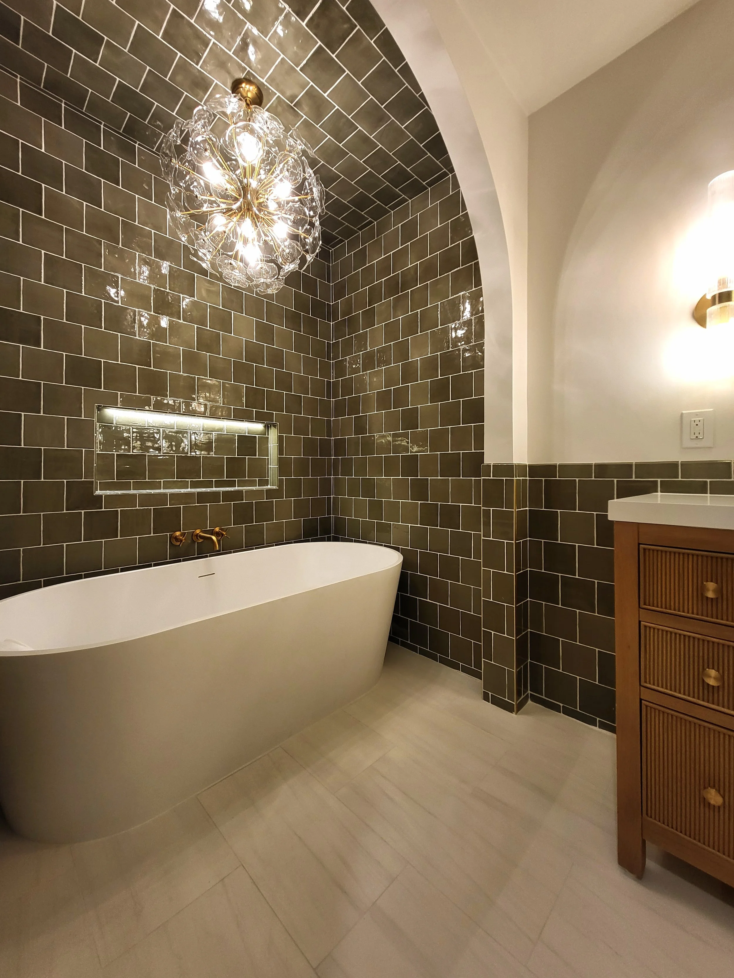 Alcove with freestanding tub, niche, and chandelier next to vanity cabinet.