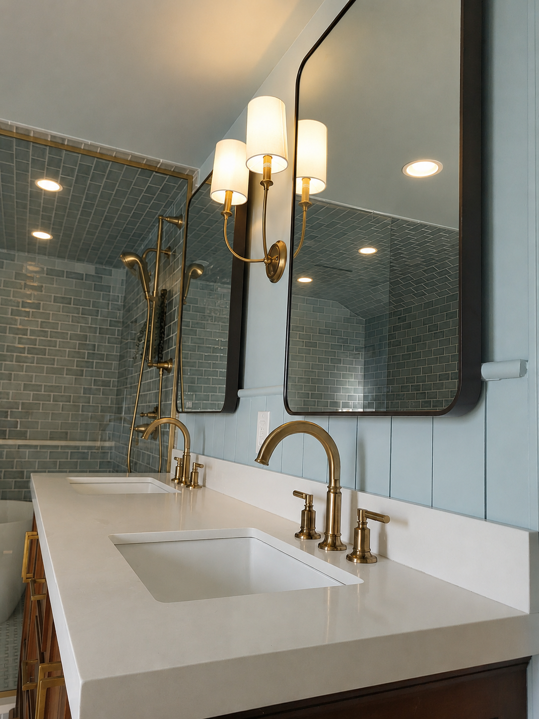 A beautiful wood vanity with marble countertop, brass faucets, mirrors, and a brass sconce with a brass shower system in the background.