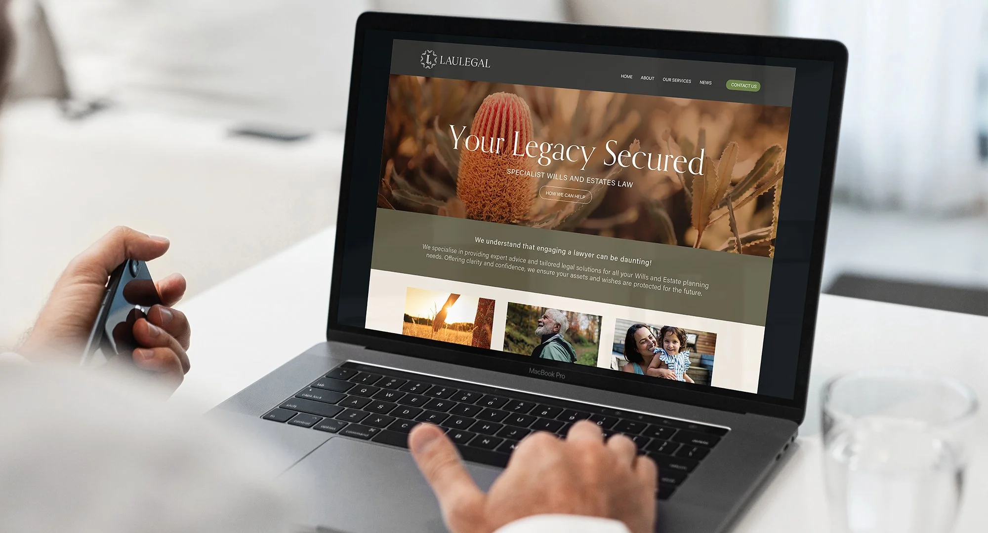 A person is holding a smartphone in their left hand while using a MacBook Pro laptop on a white desk. The laptop screen displays a legal website with the text 'Your Legacy Secured' and images of people and nature.