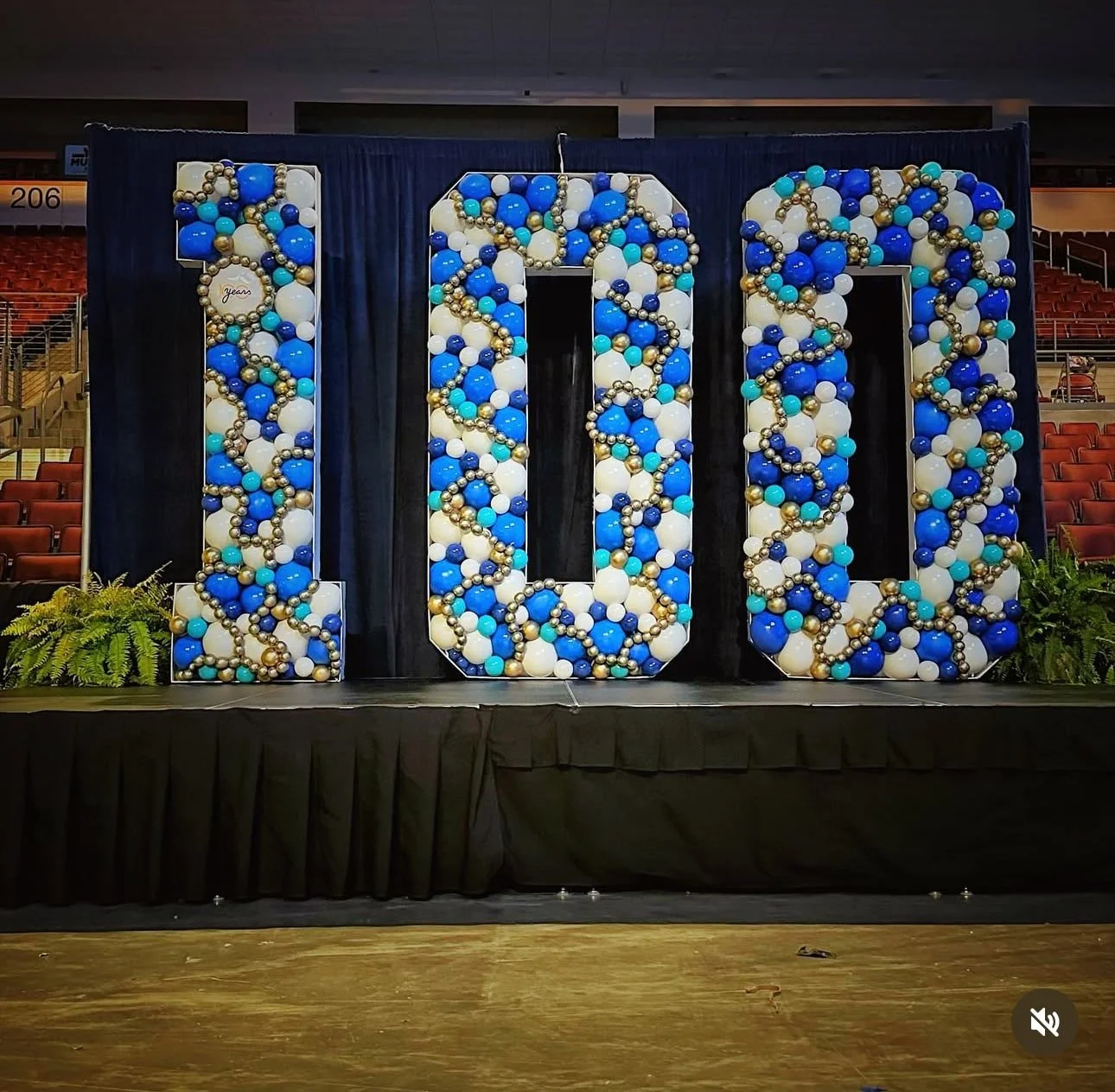 Large decorative display of the number ten made of white, blue, turquoise, and gold balloons, with a black curtain backdrop and greenery in front.