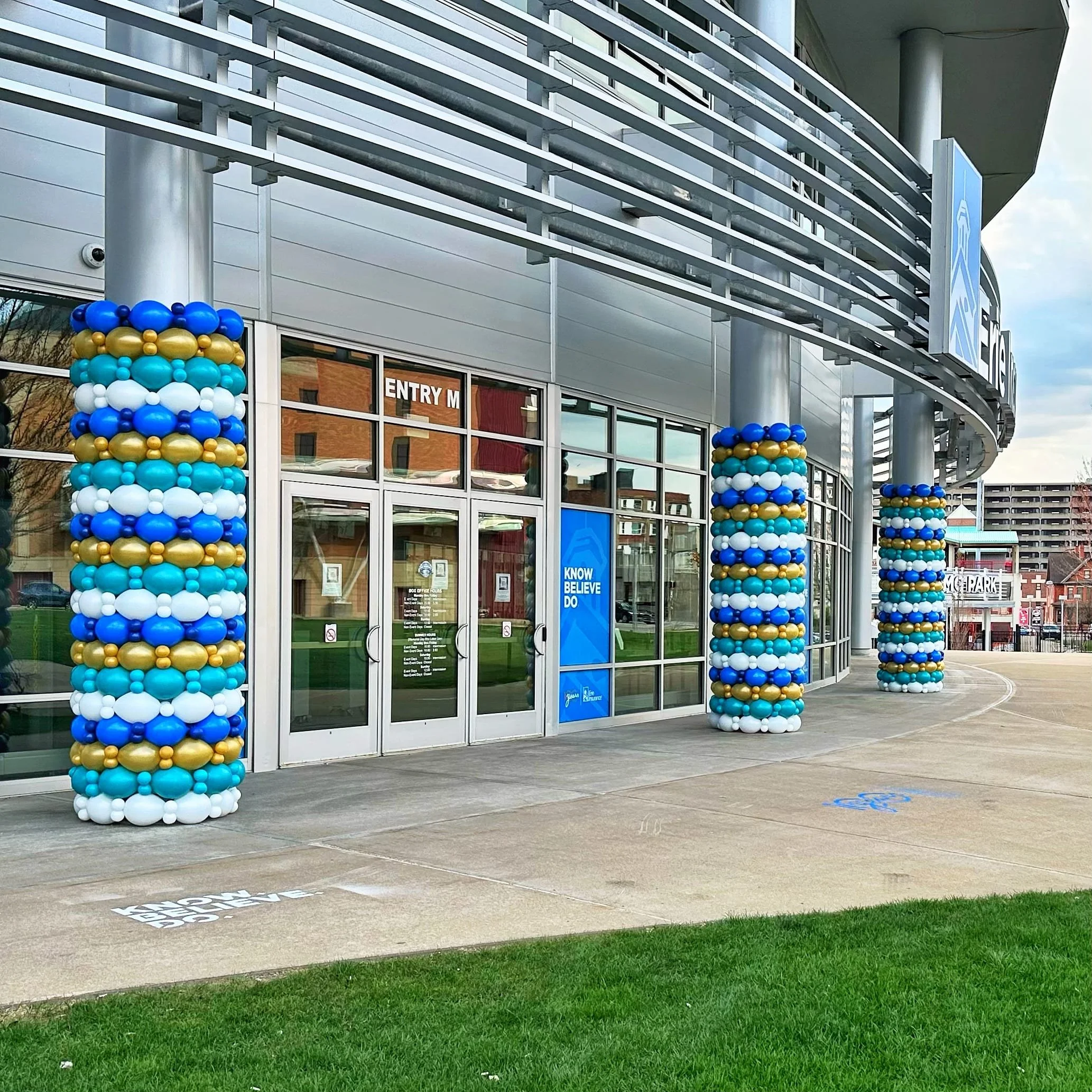 Entrance of a modern building decorated with large columns of balloons in blue, white, and gold. The building has glass doors and windows, with a blue sign that says 'Know Believe Do' and a sign indicating the entry door.