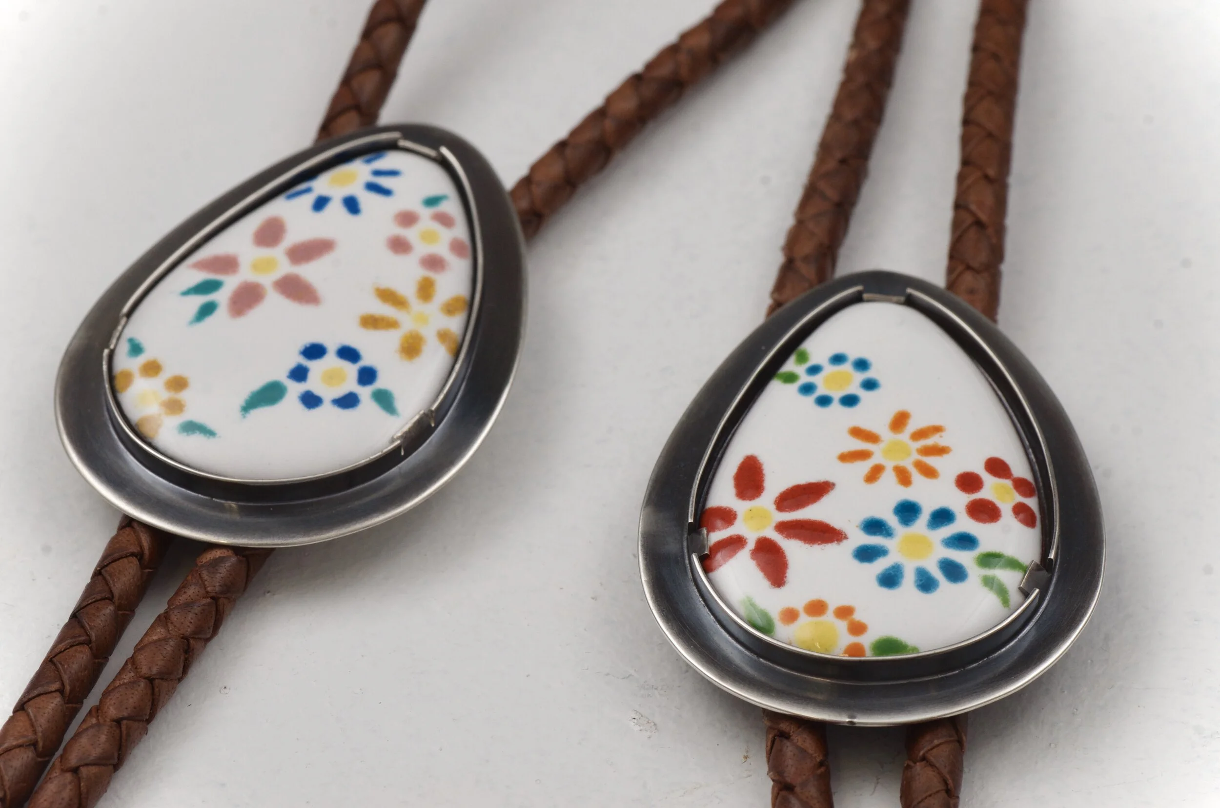 Close-up of two necklaces with teardrop-shaped pendants featuring colorful floral patterns on white backgrounds, attached to braided brown leather cords.