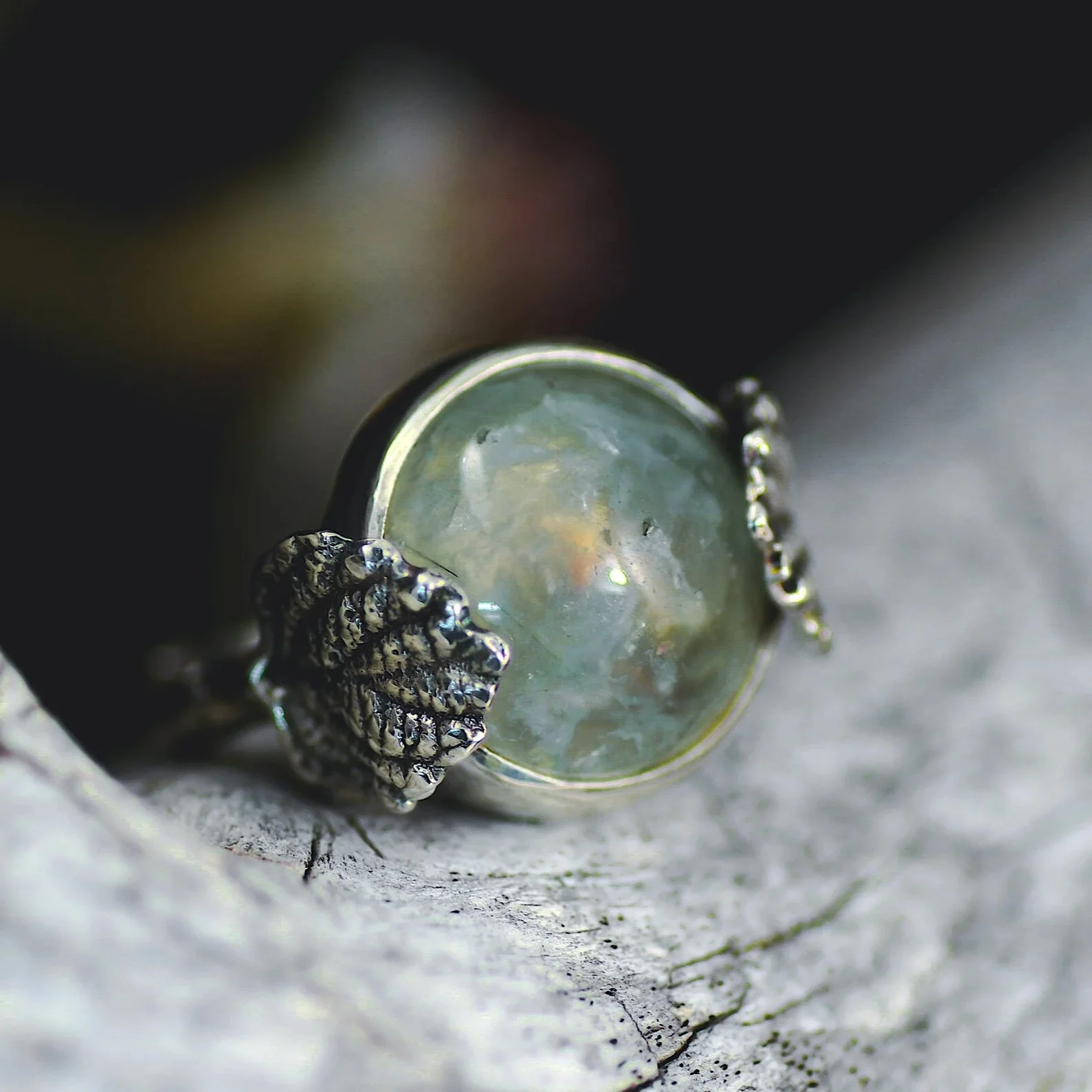 Close-up of a silver ring with a large, round, semi-translucent gemstone and textured decorative elements on a rough surface.