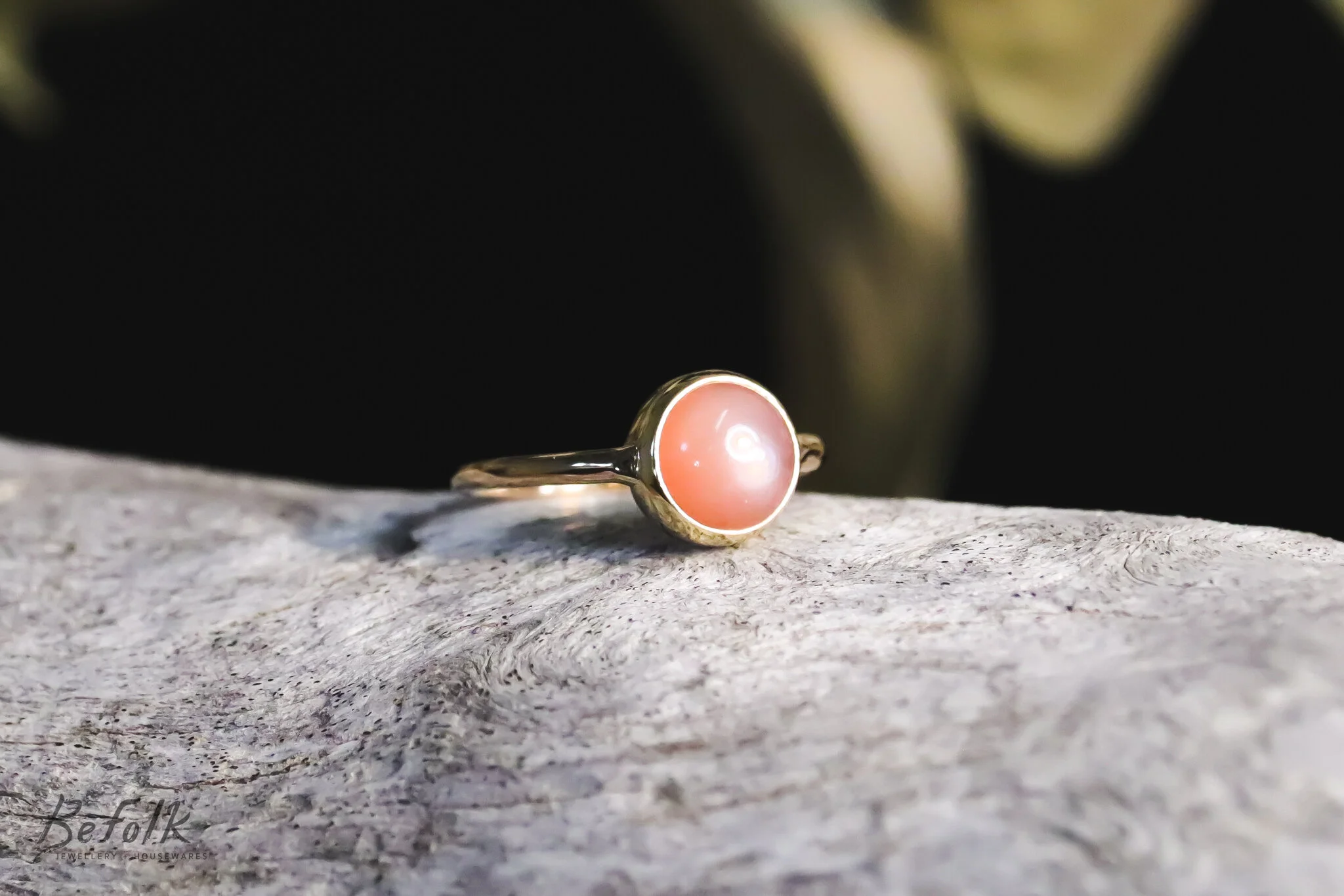 A gold ring with a pink cabochon gemstone, placed on a textured, light-colored wooden surface with a dark background.