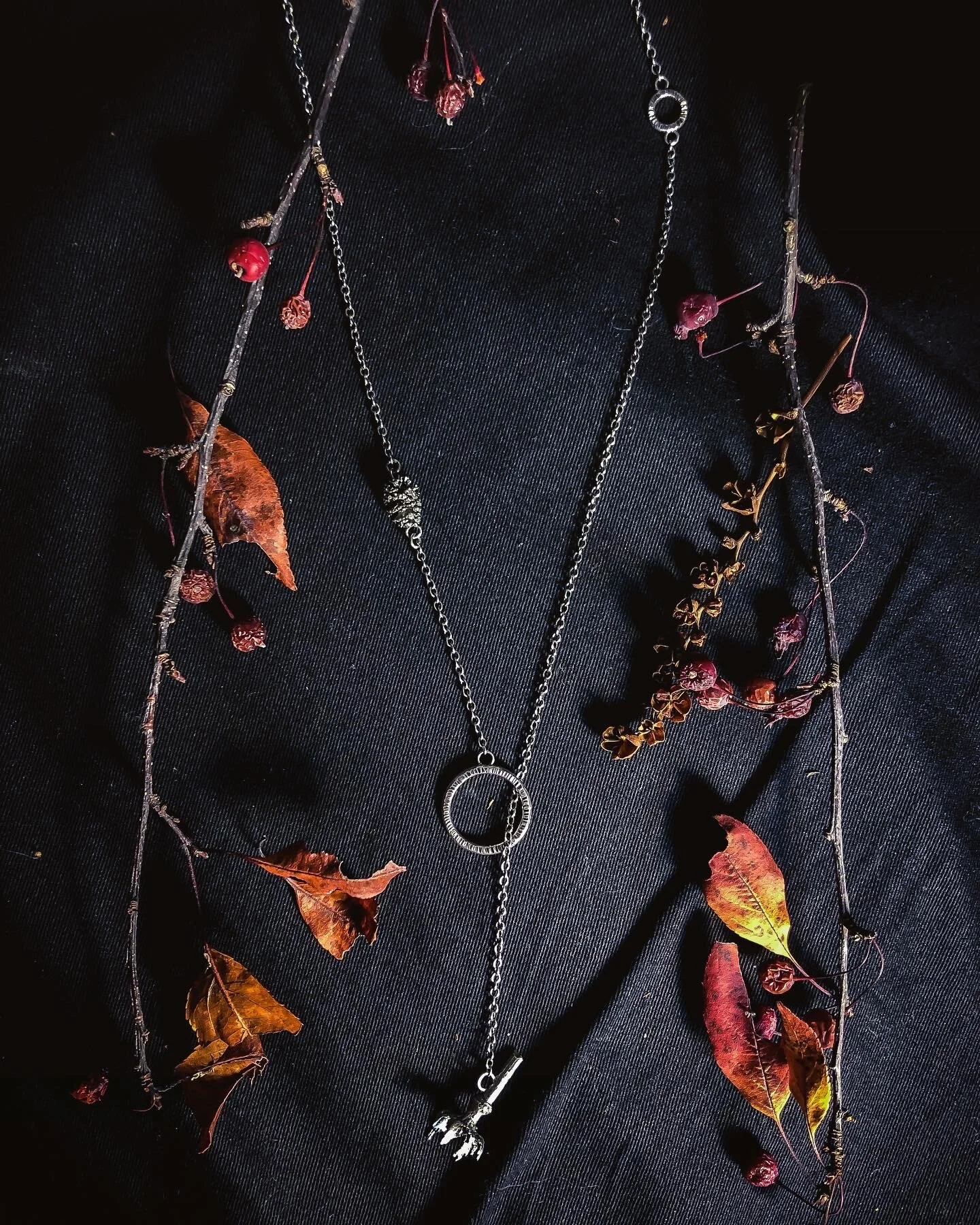 Silver necklaces displayed on a black fabric background, decorated with dried leaves, berry-like dried fruits, and small twigs.