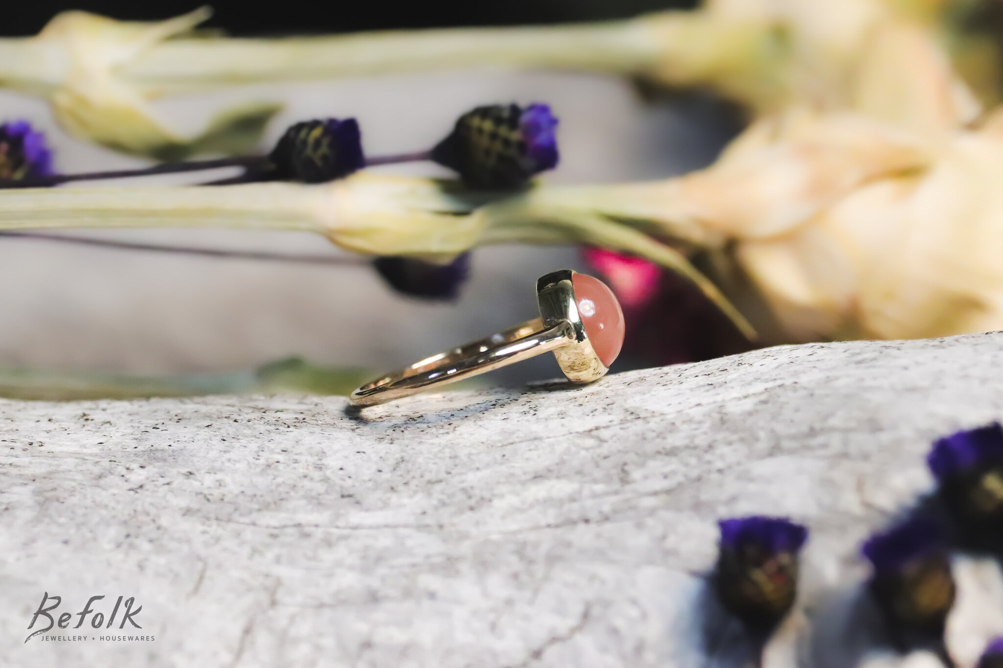 A ring with a pink gemstone on a textured white surface, surrounded by purple and cream flowers.
