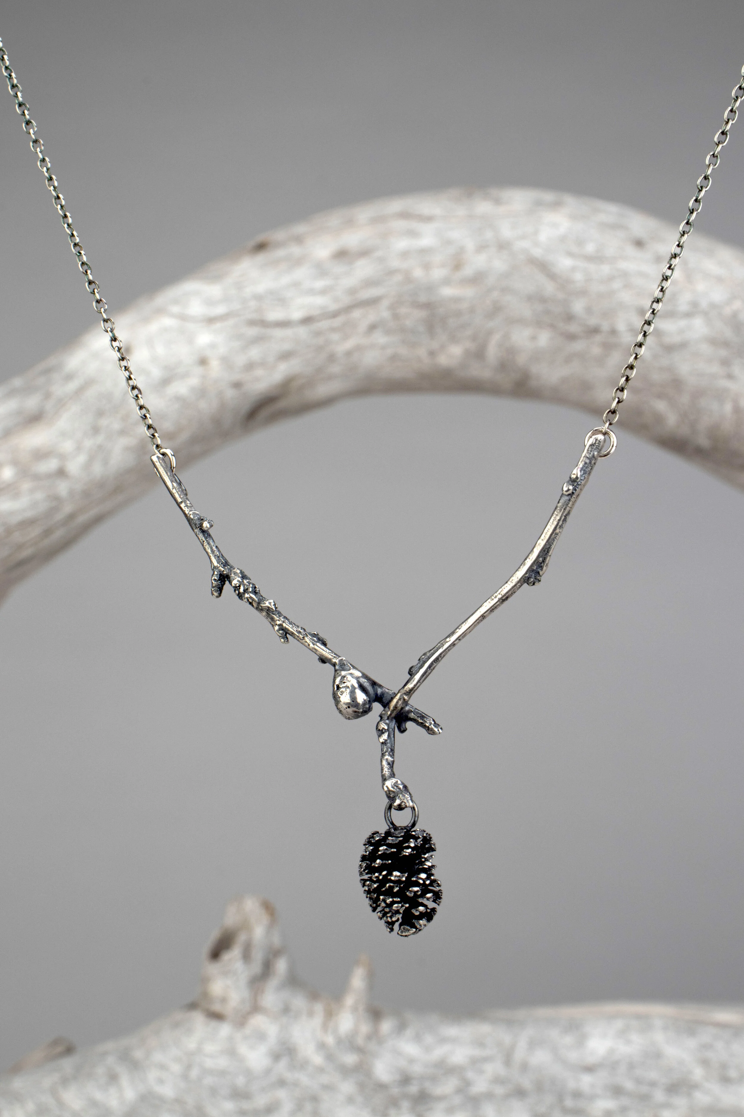 Silver necklace with a pine cone pendant hanging from a branch display against a gray background.