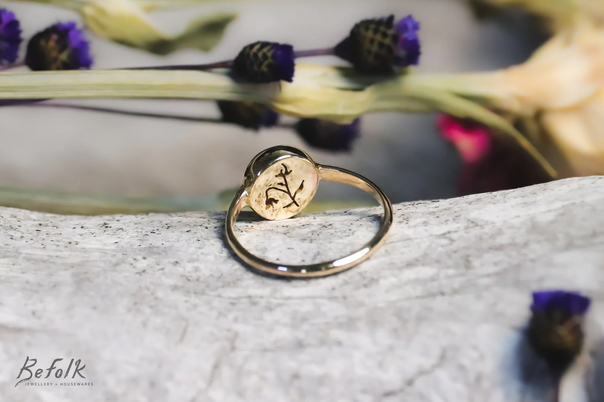 A gold ring with a round, transparent gemstone engraved with a small branch with leaves, placed on a light-colored textured surface with dried purple and pink flowers in the background.
