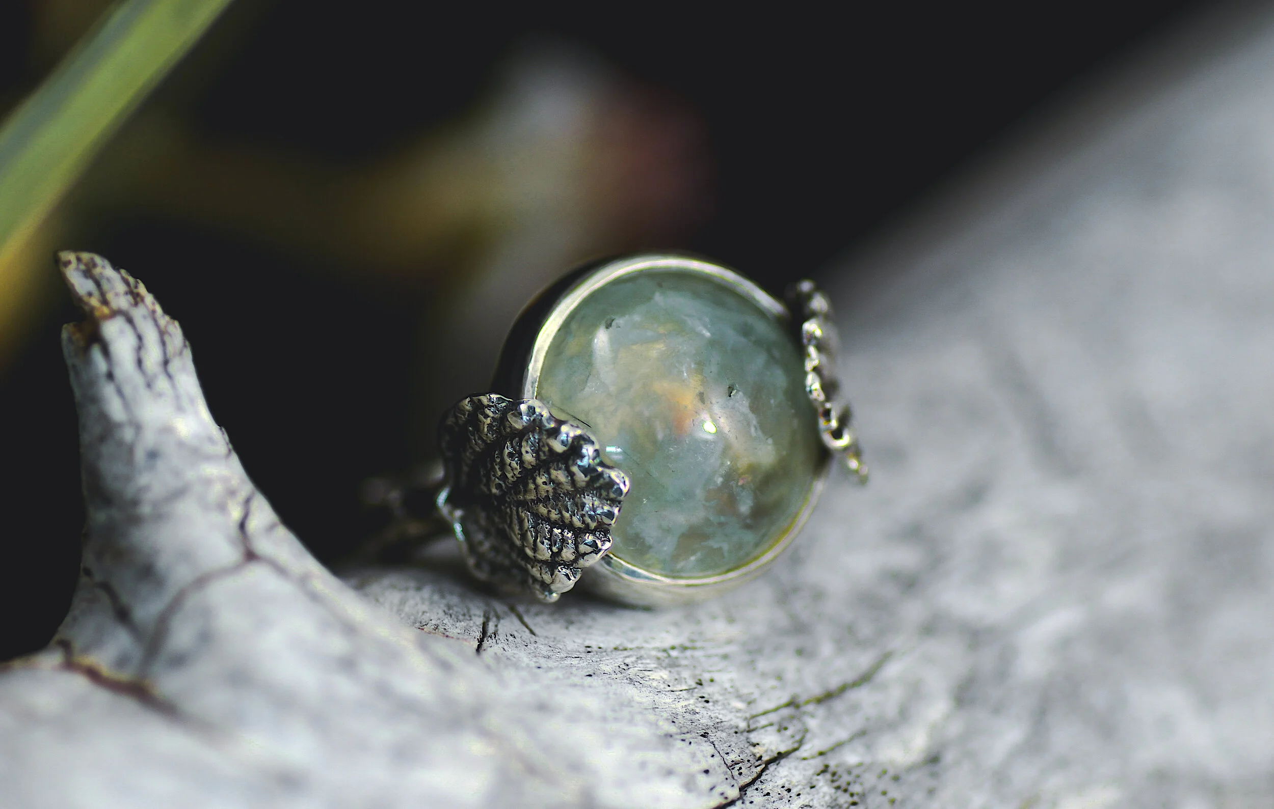 A silver ring with a round, iridescent gemstone featuring a textured leaf design on the band, placed on a weathered gray surface with some green plants in the background.