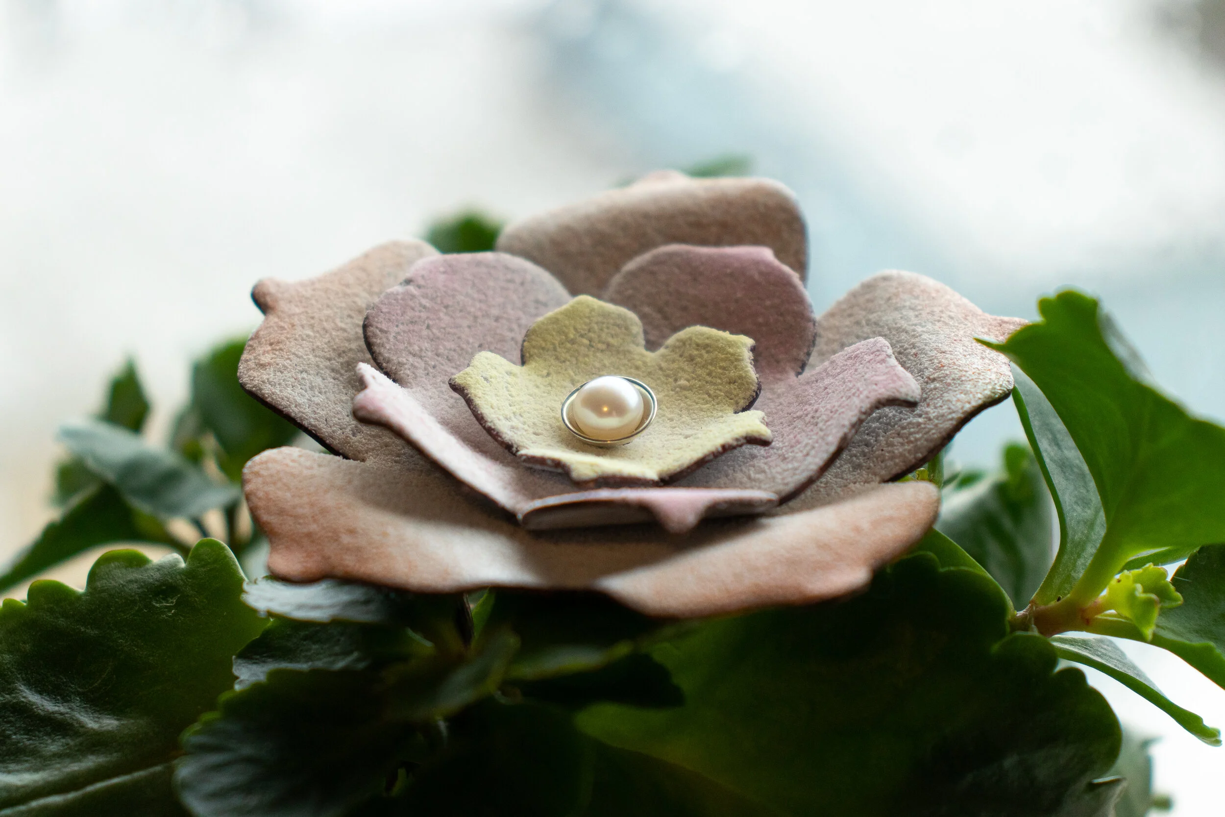 Decorative flower-shaped jewelry holder with a pearl ring on a textured pastel-colored flower, surrounded by green leaves.