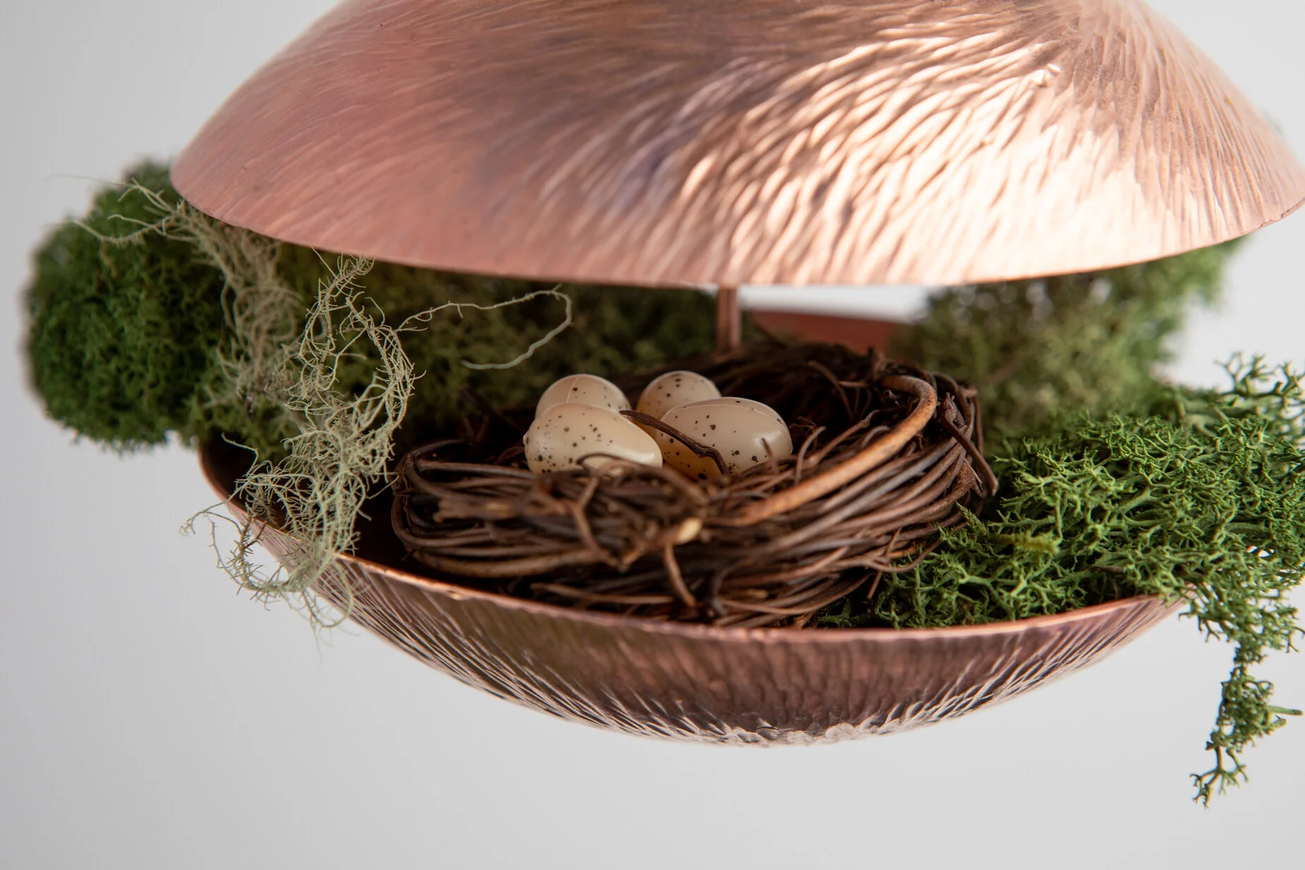 Decorative nest made of twigs with four speckled eggs inside, surrounded by moss and lichen, placed under a leaves-shaped copper lamp.