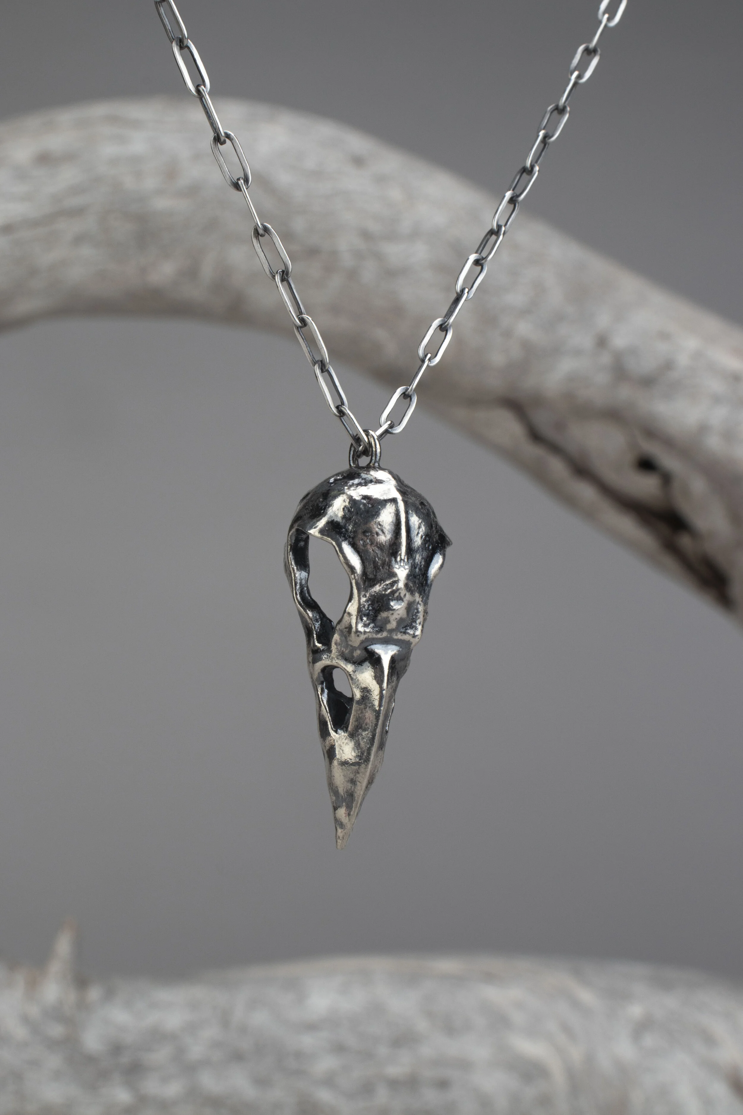 A silver necklace with a pendant shaped like a skull, hanging from a chain, displayed against a gray background with a piece of driftwood.