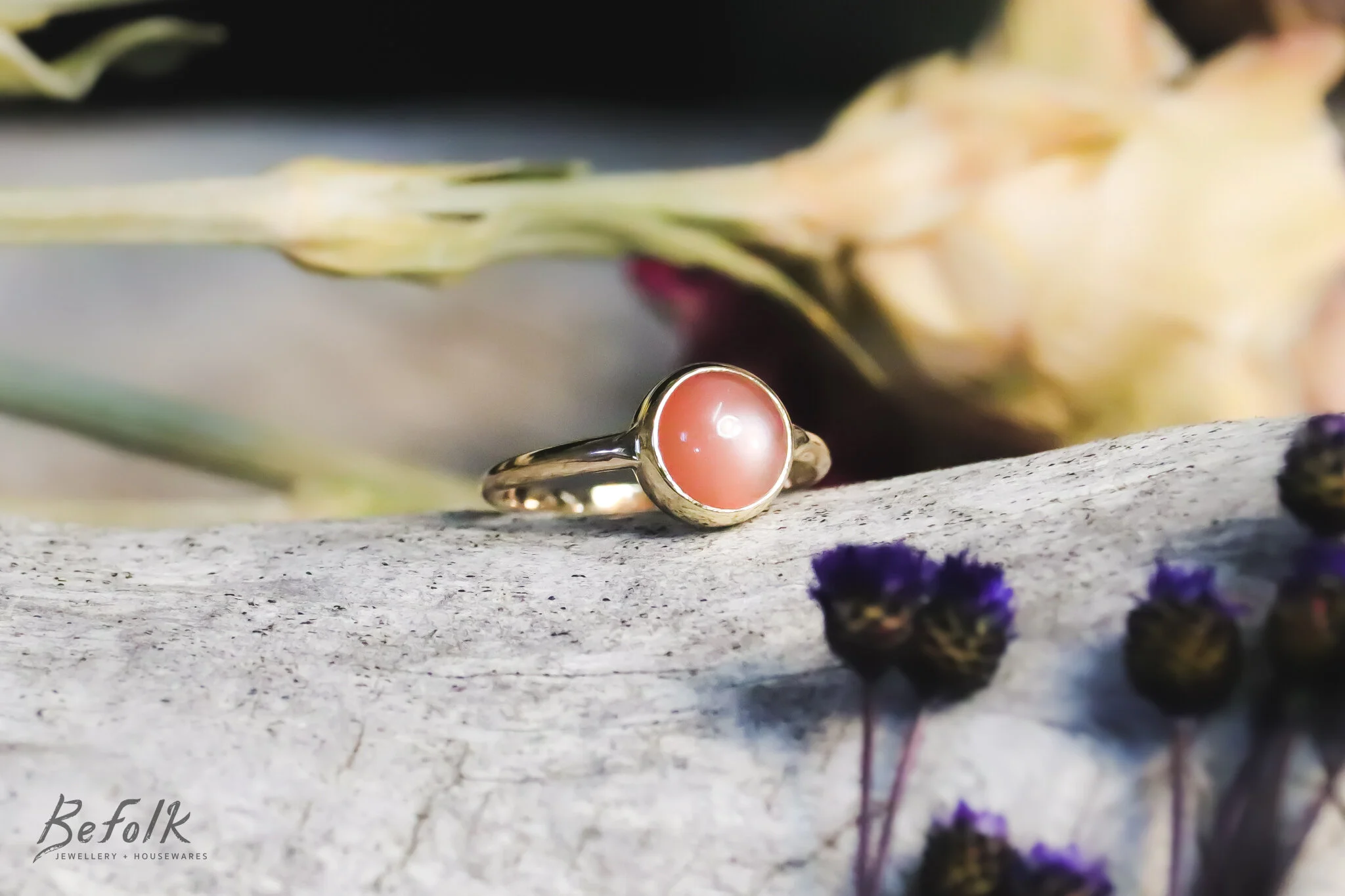 A gold ring with a pink pearl resting on a textured surface with dried purple flowers and a blurred background.