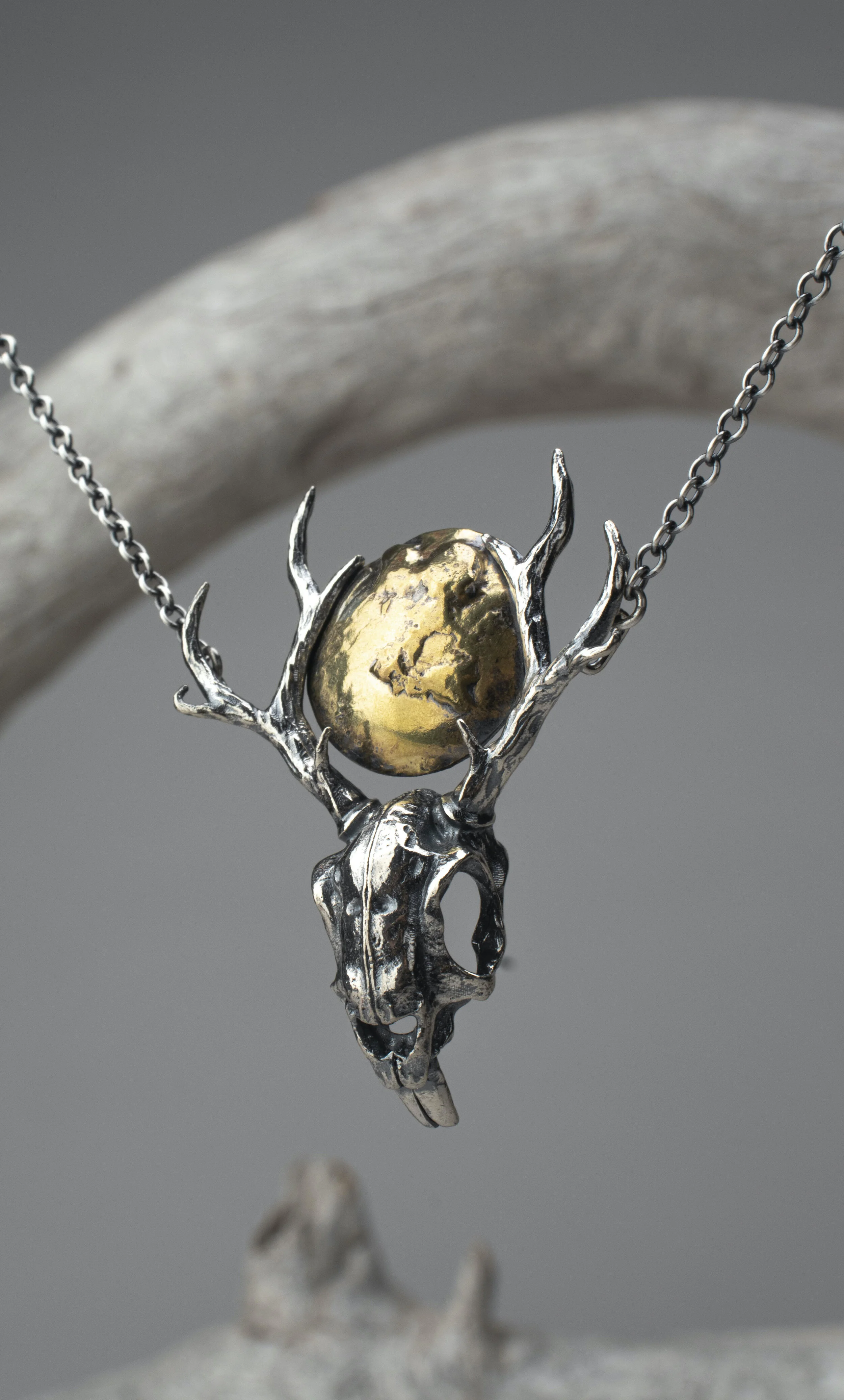 A jewelry necklace with a pendant featuring a skull and antler design, with a gold-colored sphere in the centers, set against a neutral background.