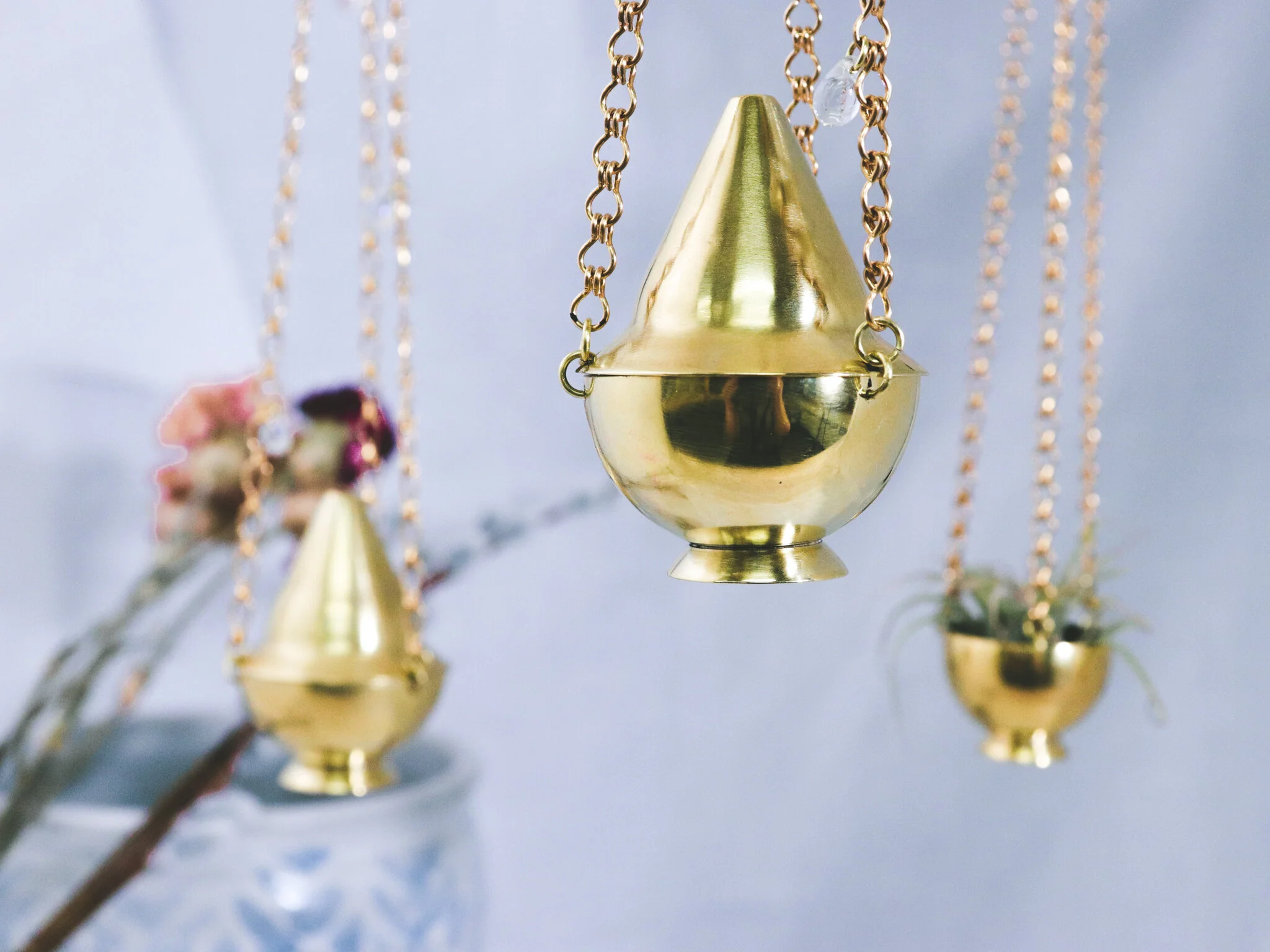 Hanged gold incense holder with other similar holders in background, hanging from golden chains with delicate plants in a glass vase, on a light-colored surface.