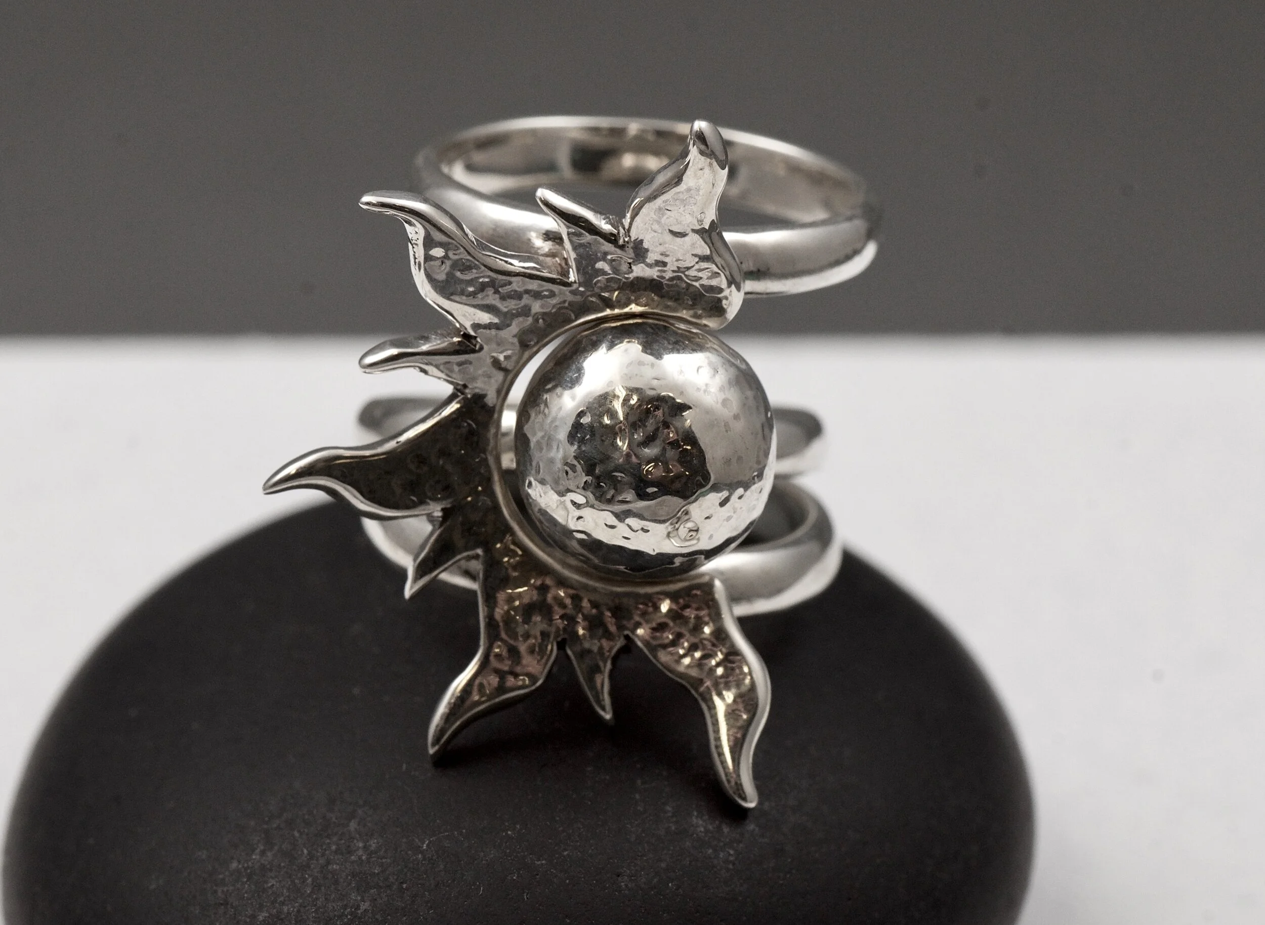 A silver ring with a sun and moon design, featuring a textured moon and a smooth sun, placed on a black surface.