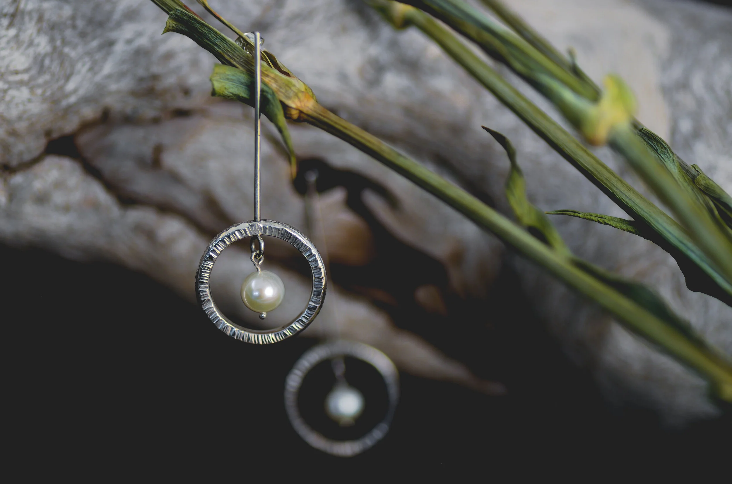 Silver earring with a pearl hanging inside a circular frame, hanging from a green plant stem against a textured background.