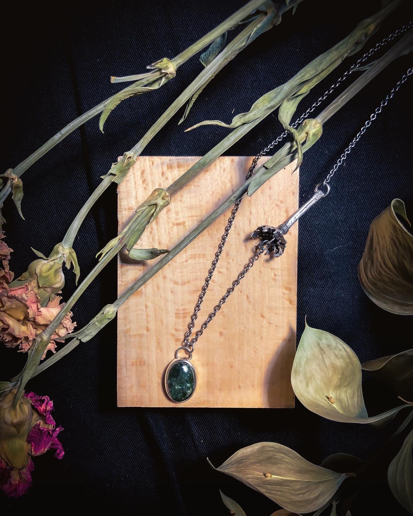 A silver necklace with a large green stone pendant placed on a wooden block, surrounded by dried flowers and leaves on a dark fabric background.