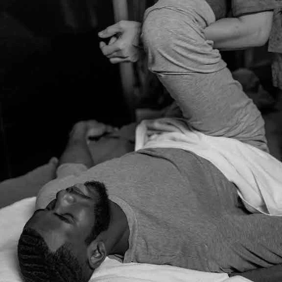 A man with a beard lies on a bed, looking relaxed, while a person in gray shorts sits on the bed nearby with their back to the camera, making a hand gesture.