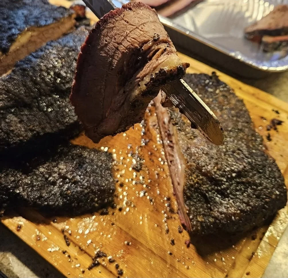 Close-up of a pitmaster demonstrating the tenderness of juicy, slow-smoked beef brisket with a rich BBQ bark at Moo Moo Smokehouse & Grill.