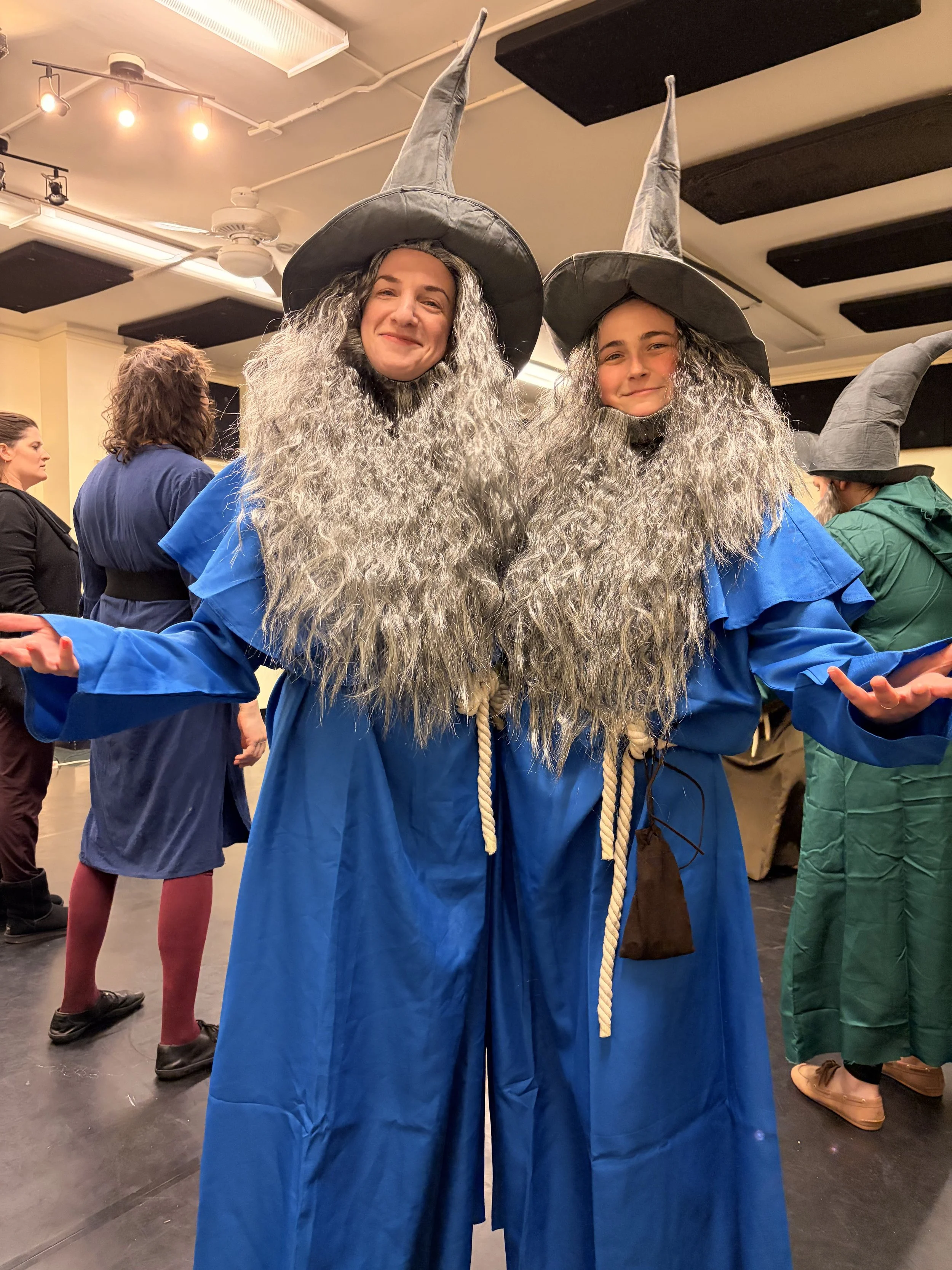 Two women dressed as wizards with long gray beards, gray pointed hats, and blue robes, standing in a rehearsal room with their arms outstretched and smiling.