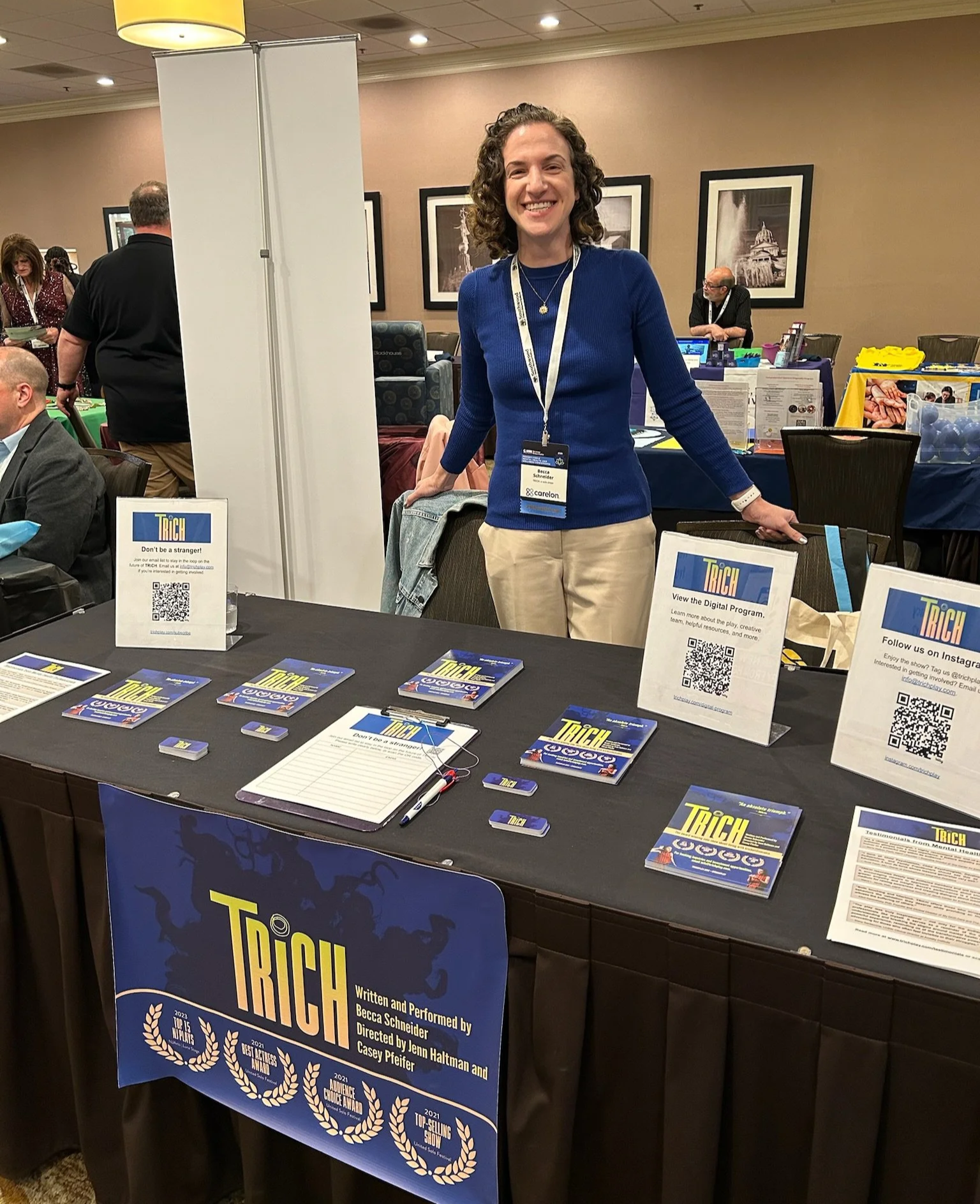 A woman standing behind a booth at an indoor event, smiling at the camera. The booth displays promotional materials for a show called 'Trich,' including flyers, QR codes, and signage. The woman wears a blue sweater, khaki pants, and a name badge.