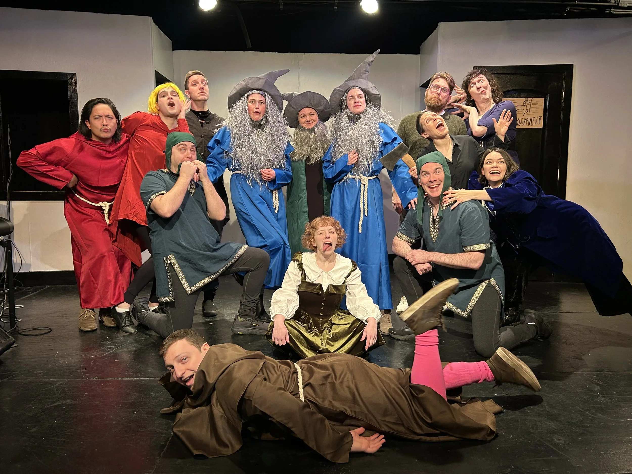 A group of actors in colorful costumes posing on stage after a theatrical performance, with some dressed as wizards with gray beards and pointy hats, others in vibrant medieval or fantasy attire, all making playful facial expressions.