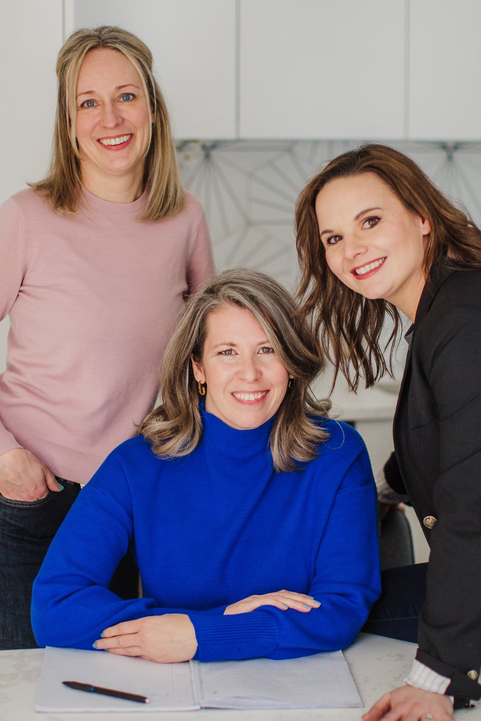 Four smiling women posing together indoors around a table with an open notebook.