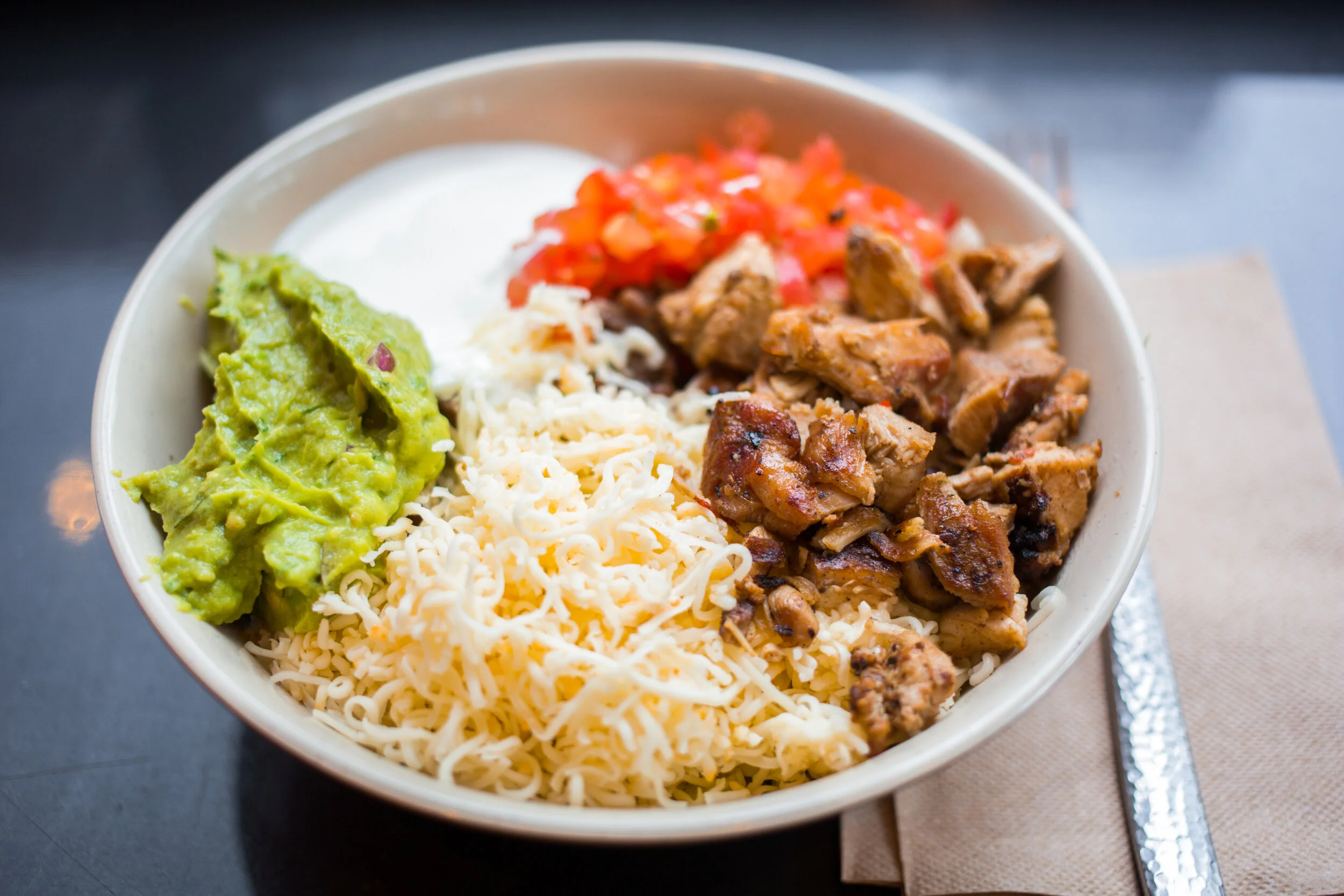 A bowl of Mexican food with shredded cheese, guacamole, grilled chicken, chopped tomatoes, and rice.