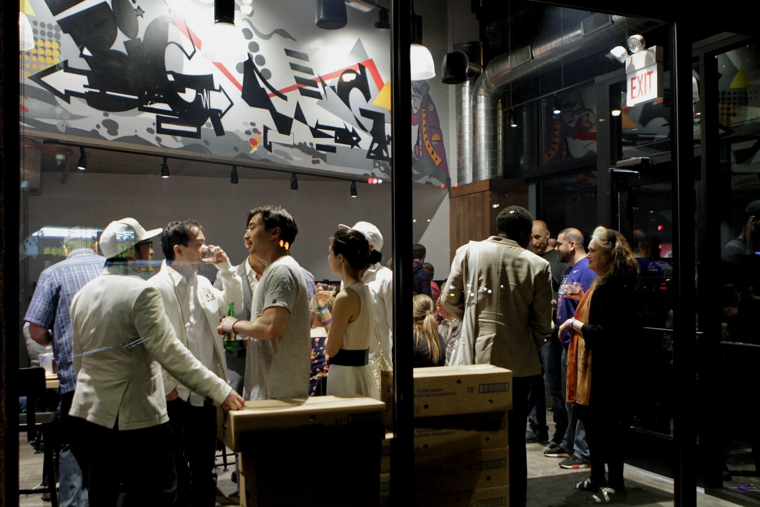 People socializing inside a modern restaurant or bar, seen through a glass window, with an artistic mural on the wall.