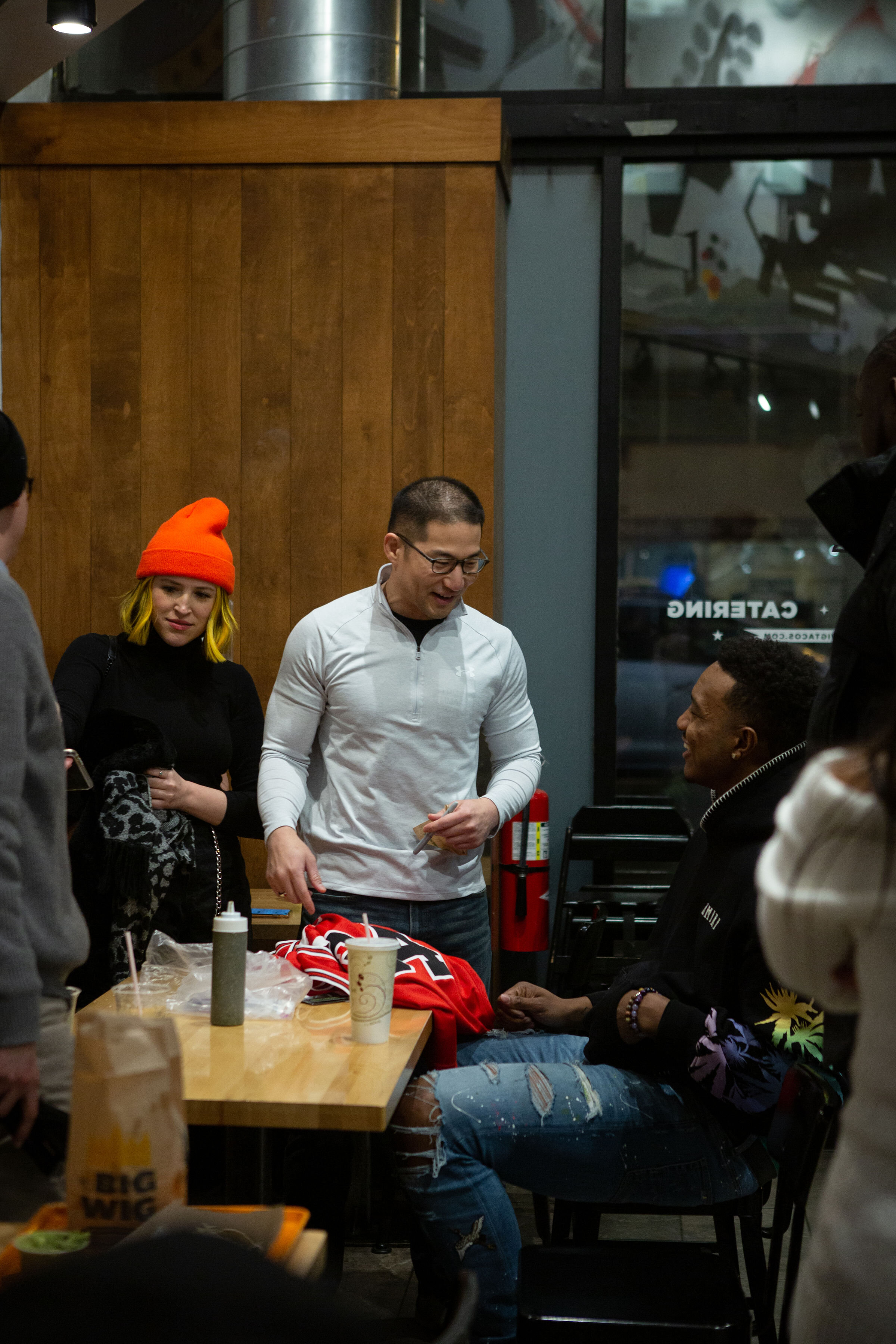 People gathered around a table in a restaurant, engaged in conversation, some smiling. A woman with yellow hair and an orange beanie stands next to a man in a white jacket, who is showing something to a seated man. The table has drinks, food, and supplies on it.
