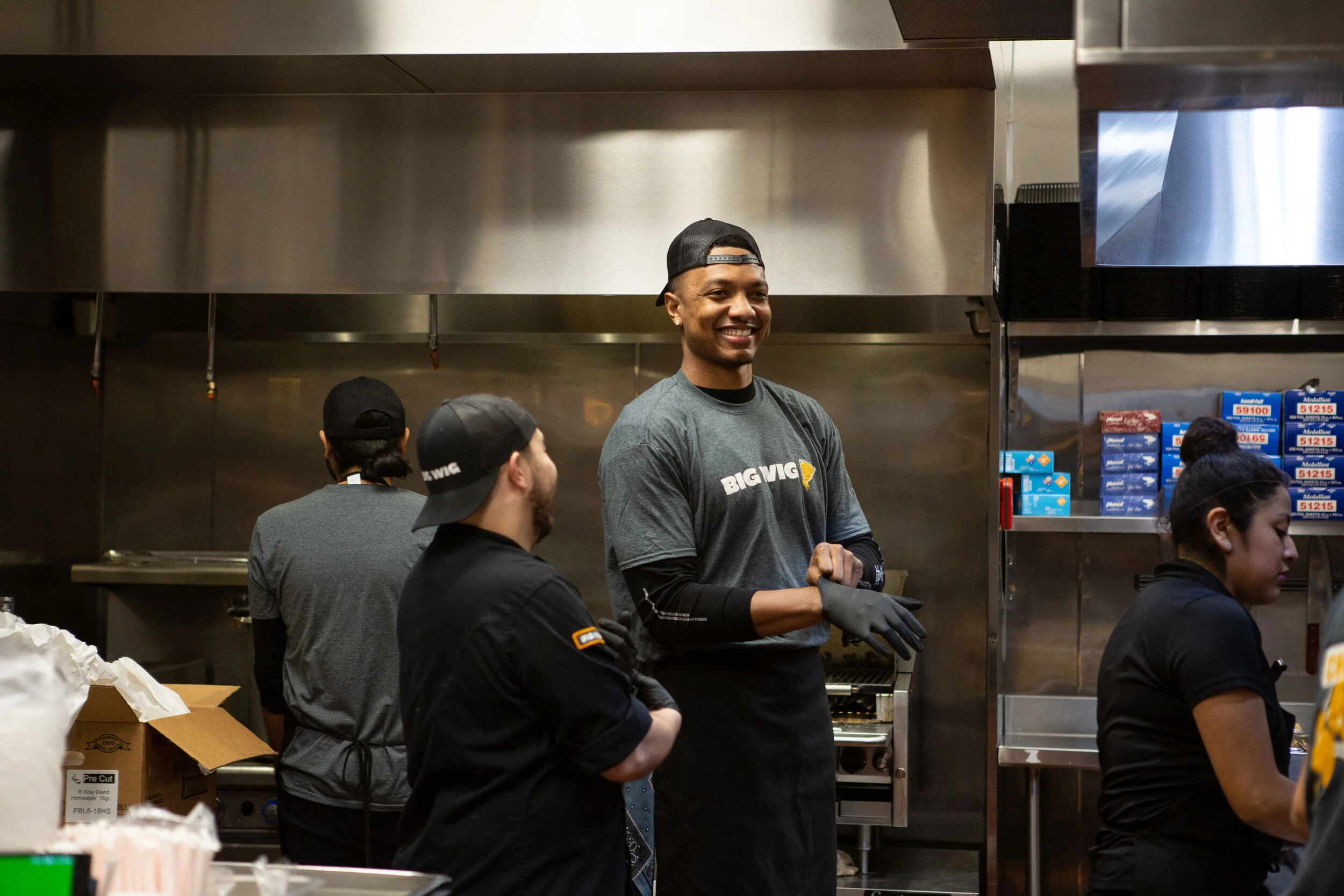 A group of people working in a commercial kitchen, with one smiling man in the center wearing a gray t-shirt and black apron, with other workers around him.