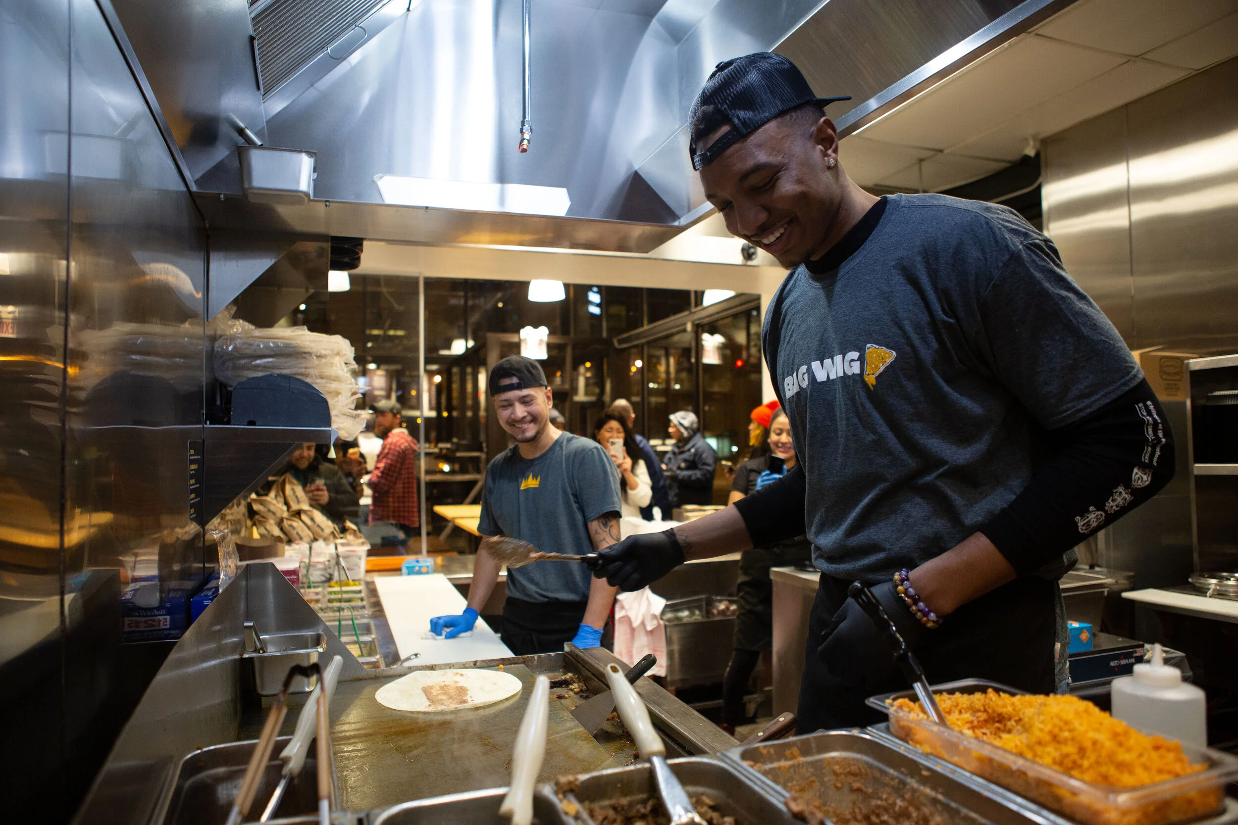 Two male workers preparing food in a restaurant kitchen with smiling customers in the background.