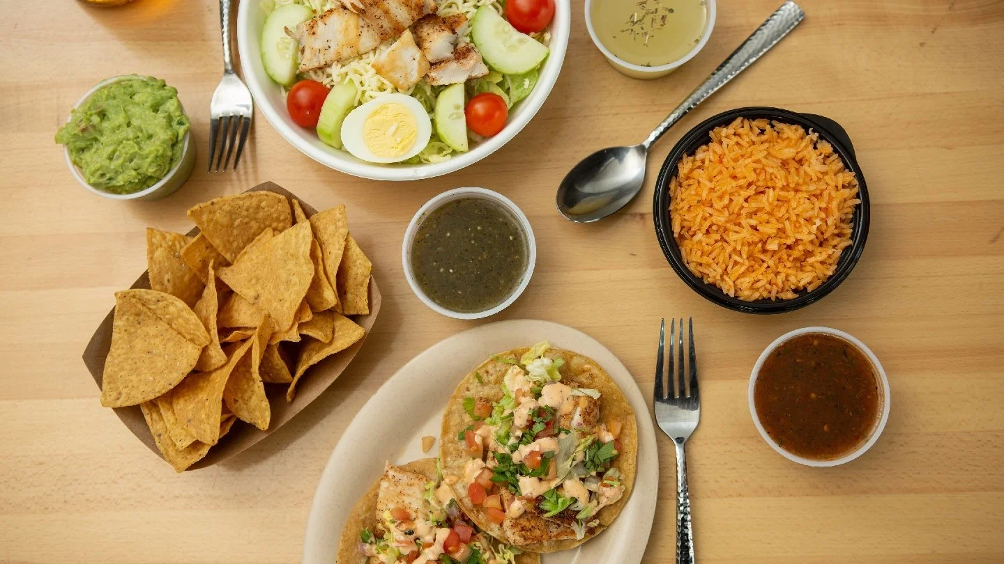 A Mexican meal with a bowl of salad, guacamole, tortilla chips, tacos with diced meat, shredded cheese, and vegetables, a bowl of Mexican rice, and several small cups of salsa and sauces on a wooden table.