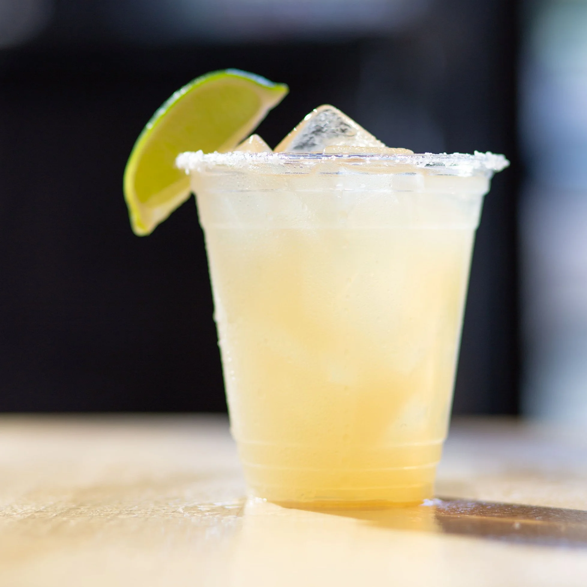 A close-up of a margarita cocktail in a clear plastic cup with salt on the rim, garnished with a lime wedge, set on a wooden surface with a blurred dark background.