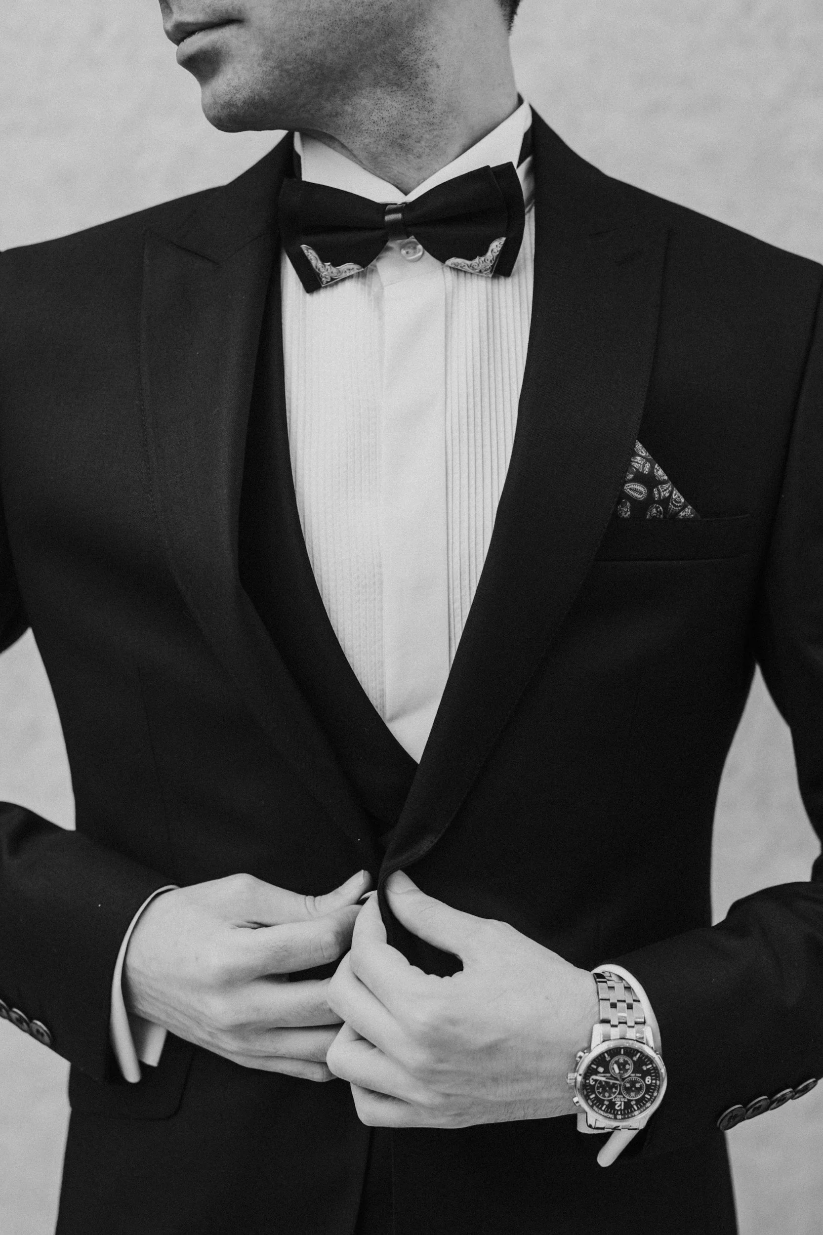 Close-up of a man in a tuxedo adjusting his suit jacket, featuring a bowtie, pocket square, and a wristwatch.