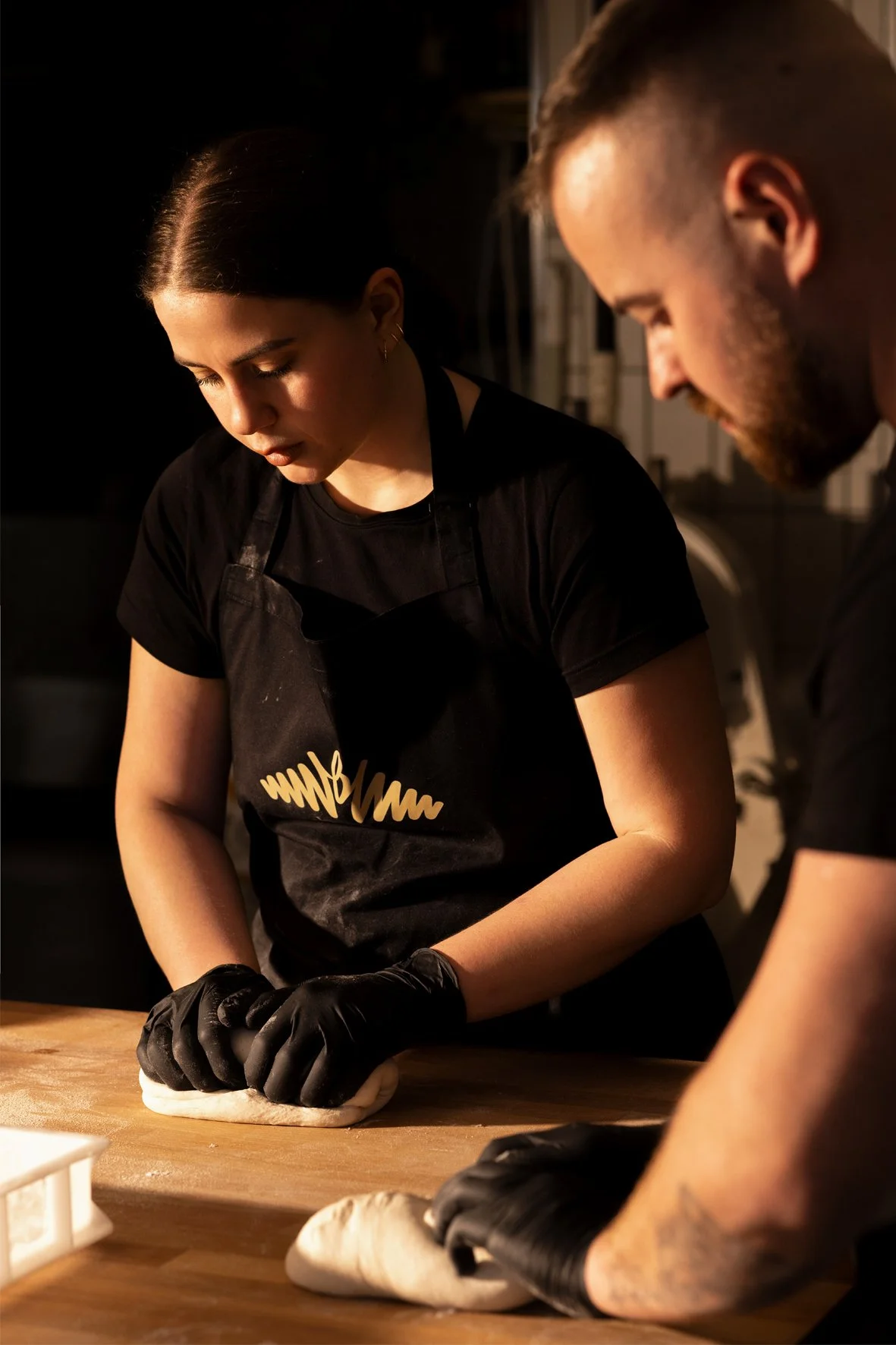 A woman and man working together on dough in a kitchen, both wearing black gloves and black shirts, with the woman wearing a black apron.