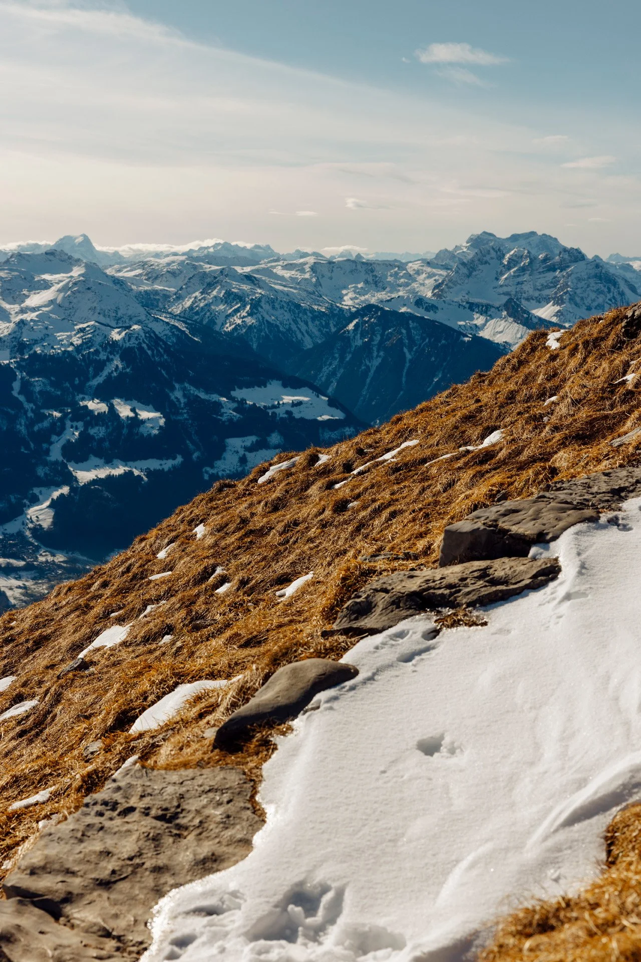 Snow-covered mountain slope with rocks and patches of brown grass, overlooking a vast mountain range under a cloudy sky.