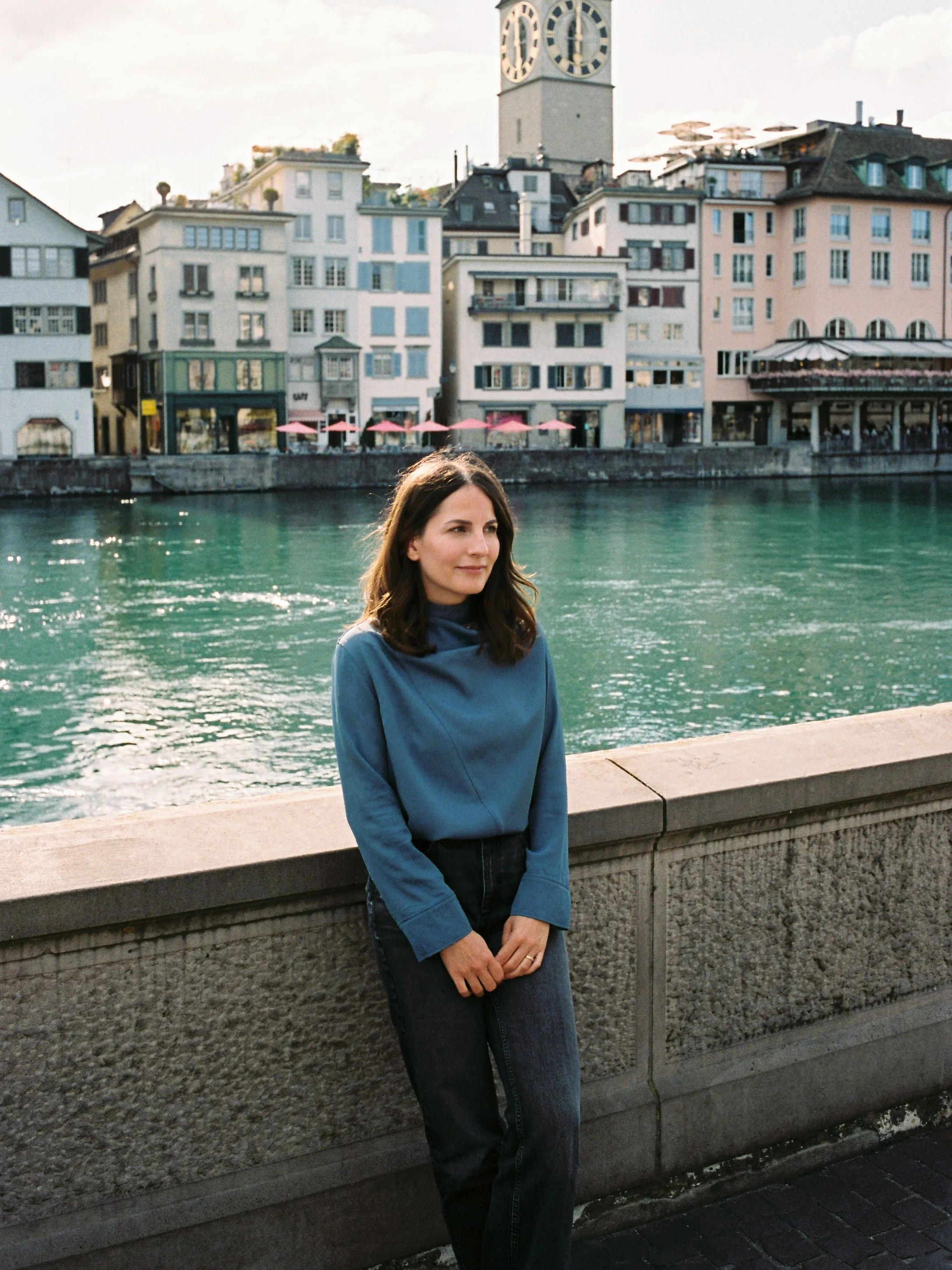 A woman with shoulder-length dark hair, wearing a blue long-sleeve top and dark jeans, standing outdoors by a stone railing along a body of water with colorful buildings and a clock tower in the background.