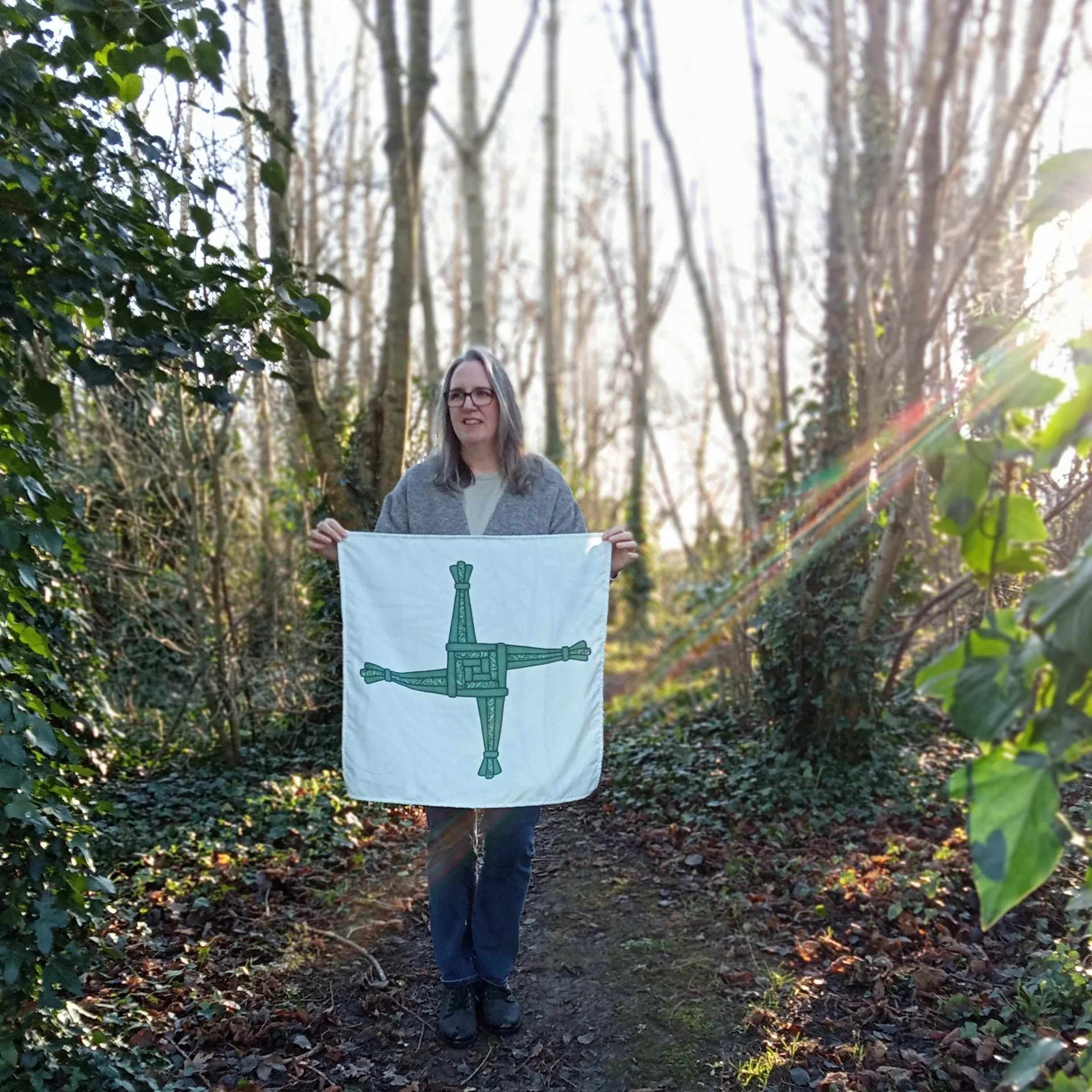 Woman standing on a narrow woodland path holding a Brat Bhríde cloth with a green St Brigid’s cross design, surrounded by bare trees and ivy.