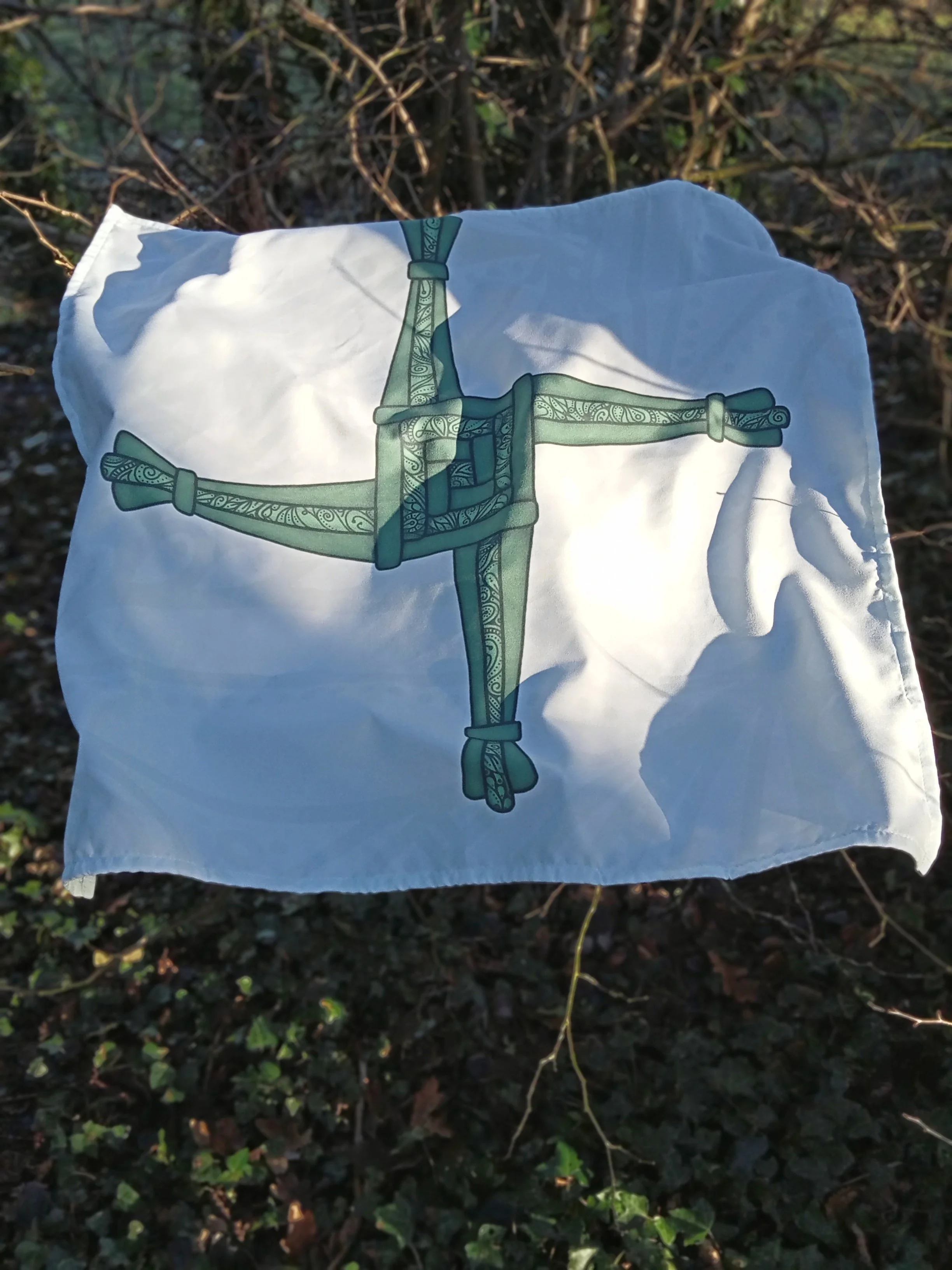 Brat Bhríde cloth laid out outdoors in woodland, featuring a green St Brigid’s cross design on white fabric with sunlight and shadows across it.