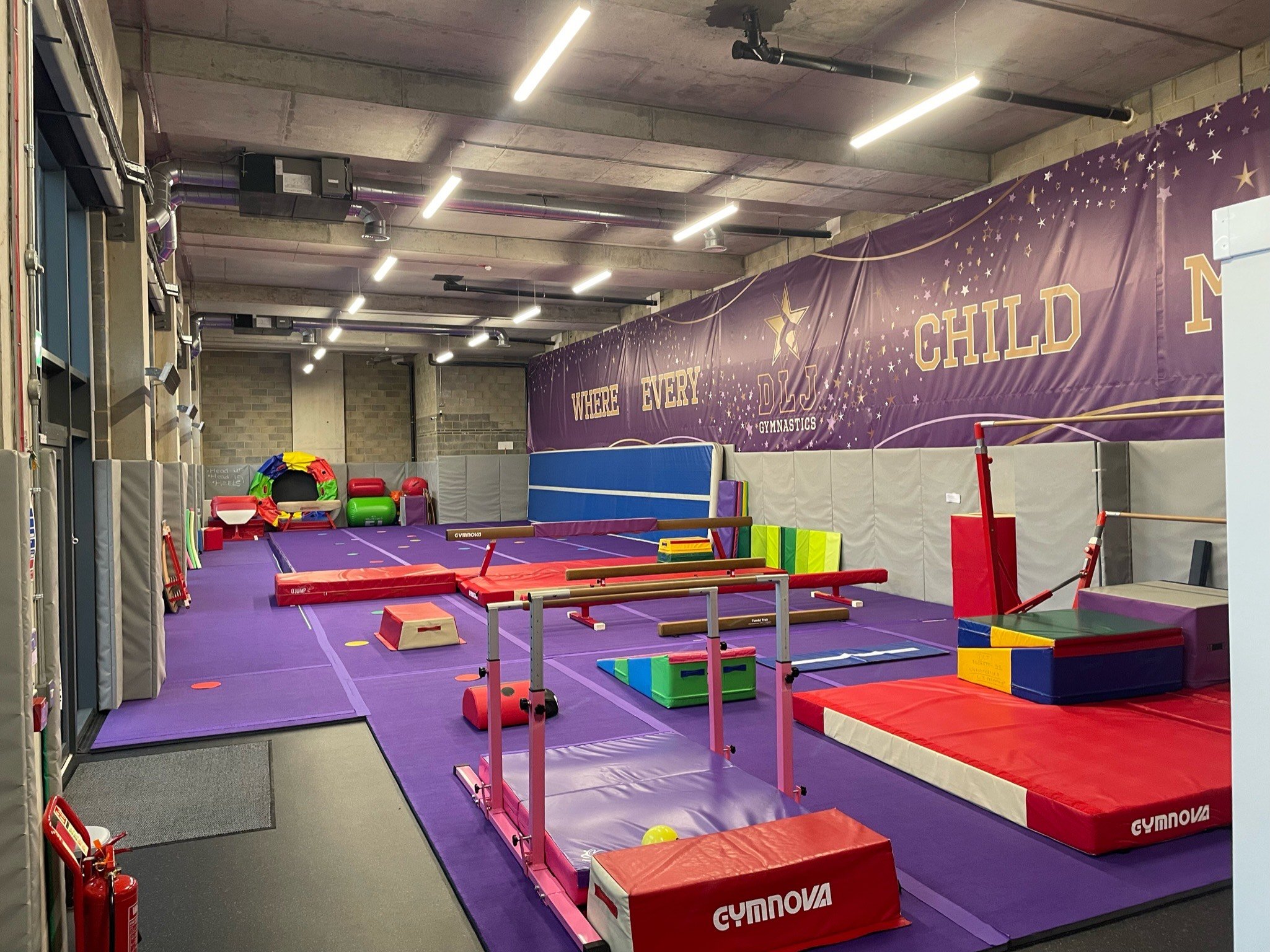 Indoor gymnastics training facility with purple mats, various colorful gym equipment like balance beams, vaulting platforms, and soft blocks, with a motivational banner on the wall.