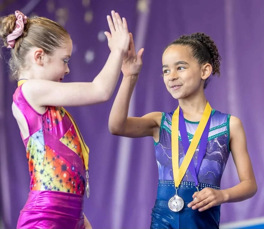 Group of young girls in purple and black gymnastics leotards practicing on the uneven bars in a gym with a purple backdrop that says 'MATTERS' and decorated with silver tinsel and snowflakes.