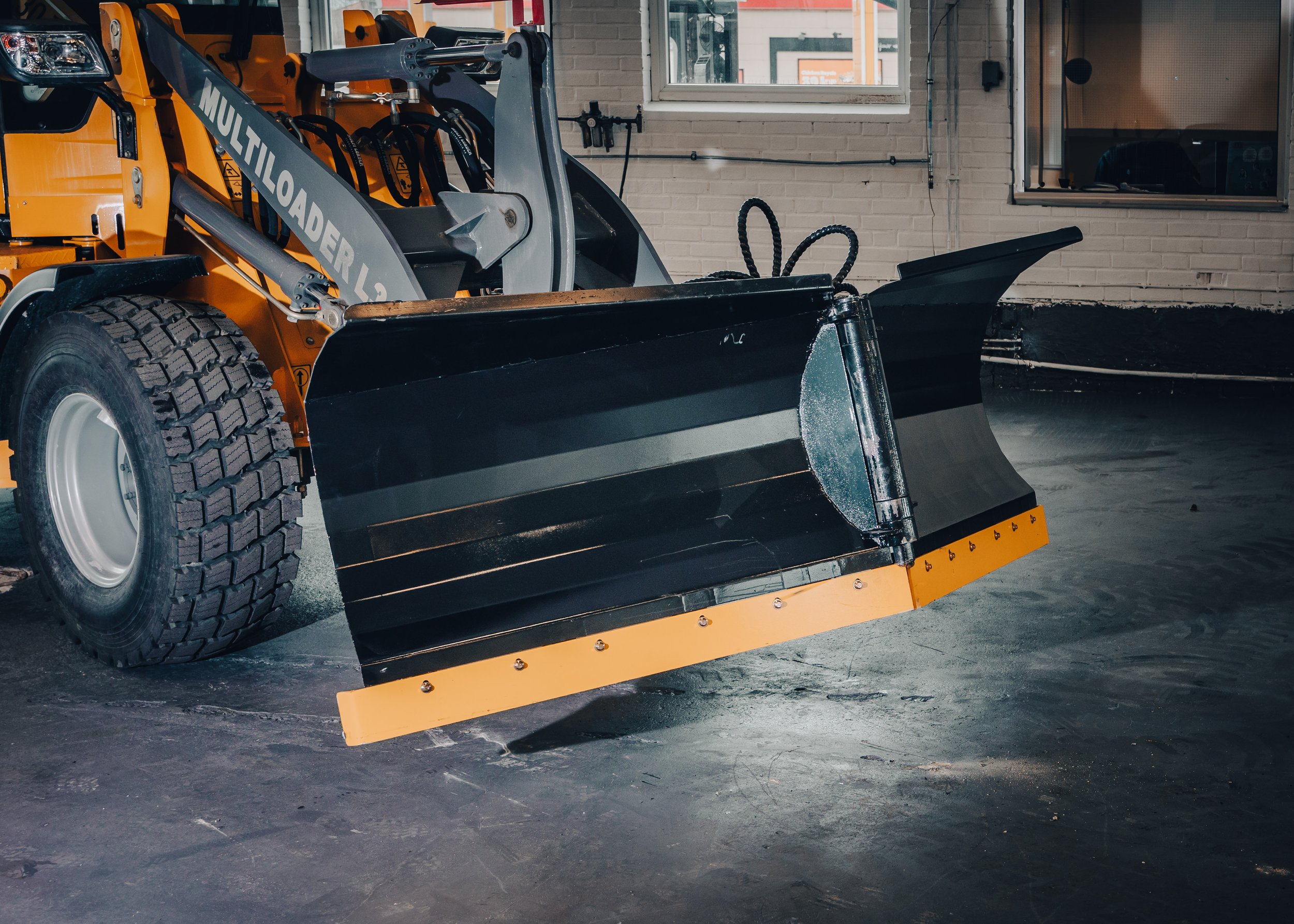 Close-up of a construction or industrial vehicle with a large black snow plow attachment at the front, parked indoors on a concrete floor.