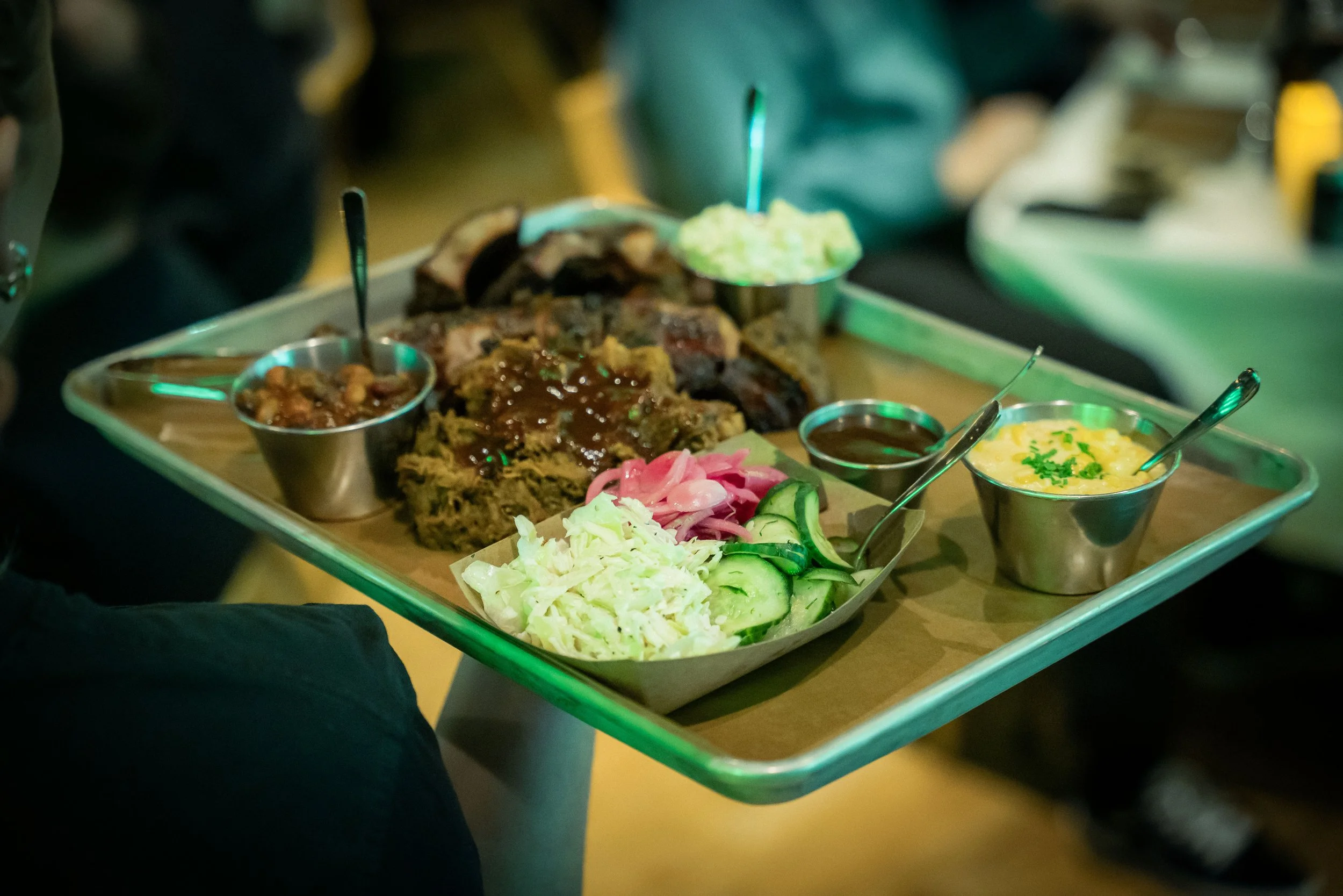 Metal tray with assorted barbecue meats, coleslaw, sliced cucumbers, pickled red onions, and small bowls of dipping sauces and sides at a restaurant.