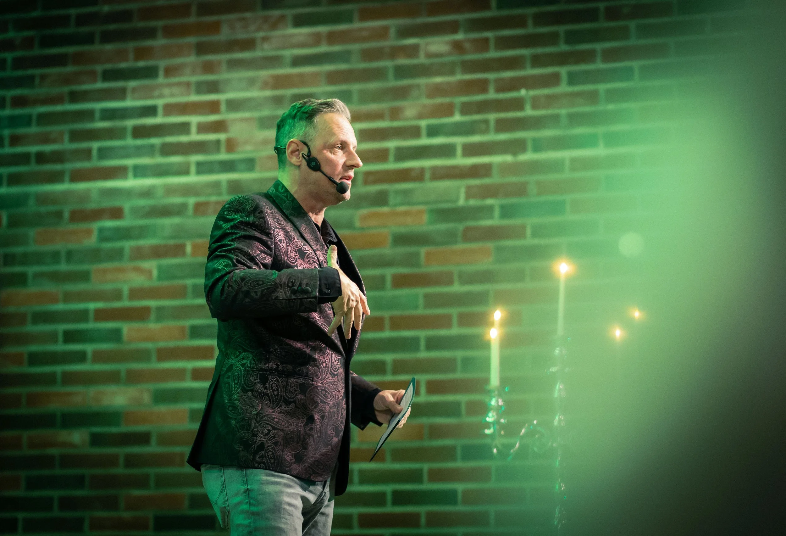 A man wearing a patterned blazer and light-colored pants giving a presentation or speech with a headset microphone, standing next to a candle holder with lit candles against a brick wall.