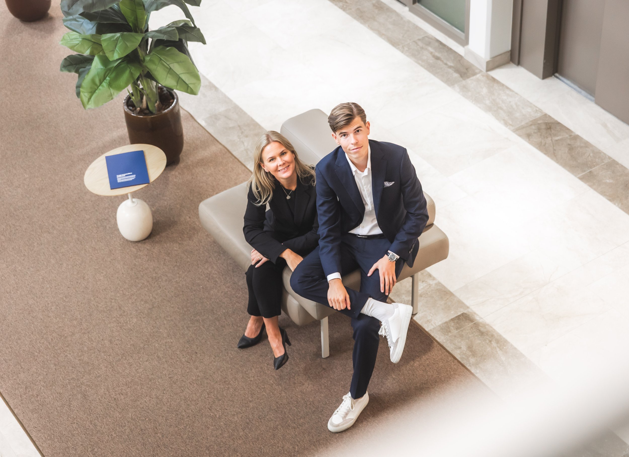 A woman and a young man sitting on a bench in a modern lobby, looking up at the camera. The woman is dressed in black with heels, and the young man is in a navy suit with white sneakers. There is a large green plant in a pot and a side table with a b
