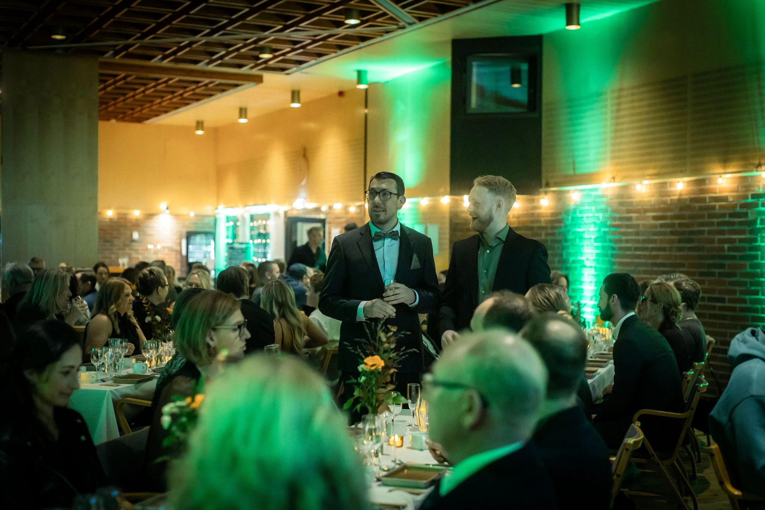 A formal banquet or dinner event with many people seated at decorated tables. Two men in suits, one wearing a bow tie, are standing and conversing, holding glasses. The room has warm lighting, green accent lights on the walls, and a brick wall backgr