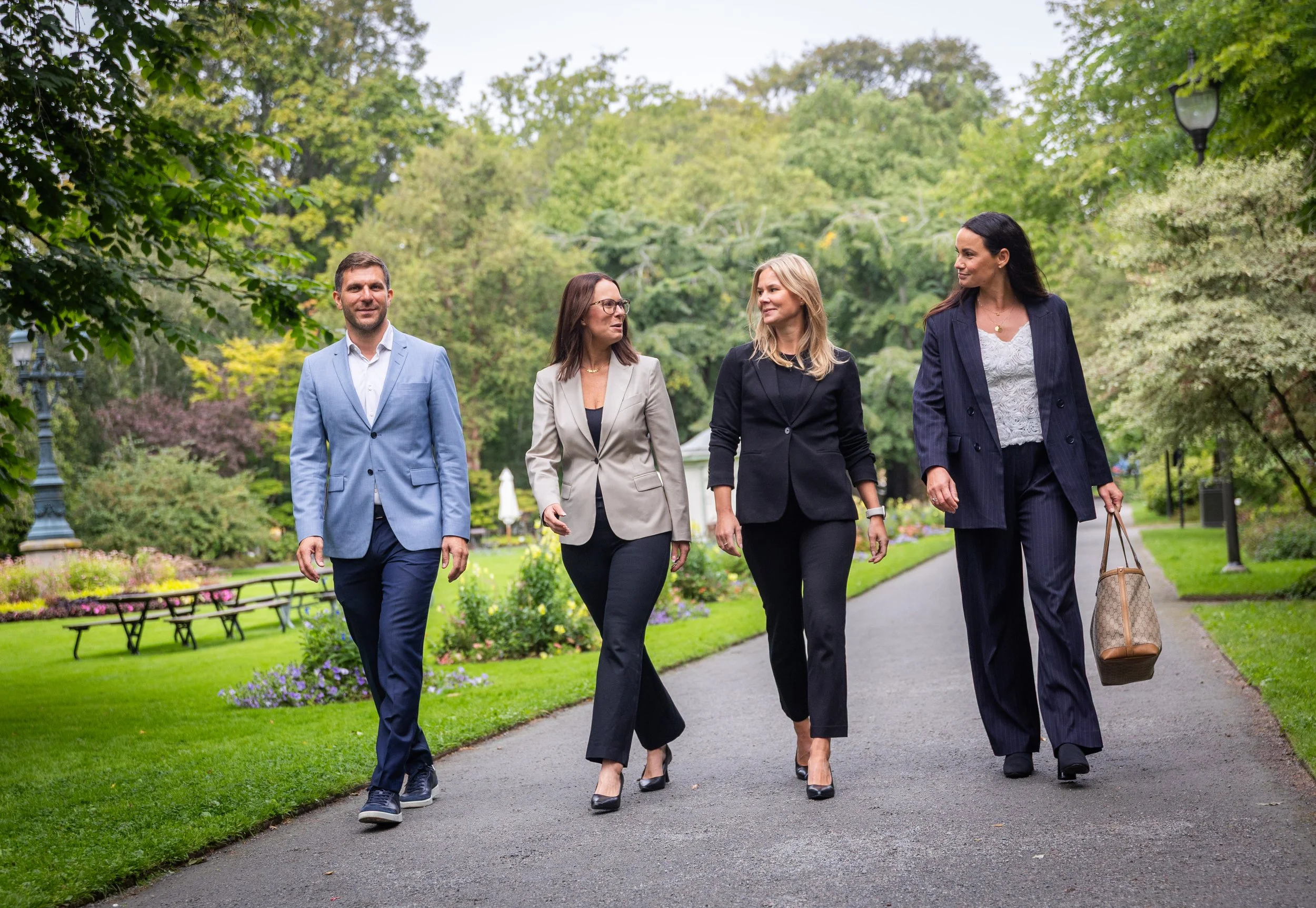 Four professionally dressed people walking together in a park with green trees and flowers.